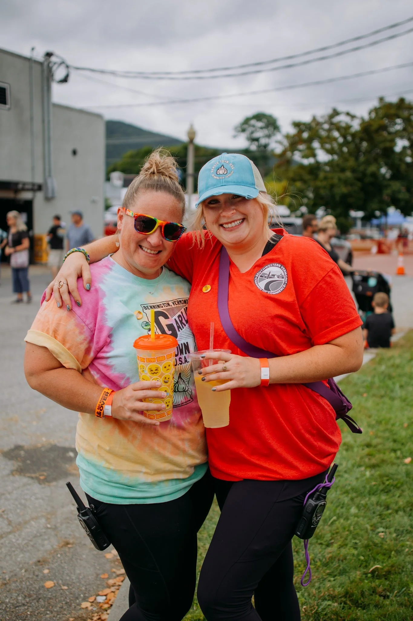Two women smiling and posing together at an outdoor event, holding drinks, with others and tents visible in the background.
