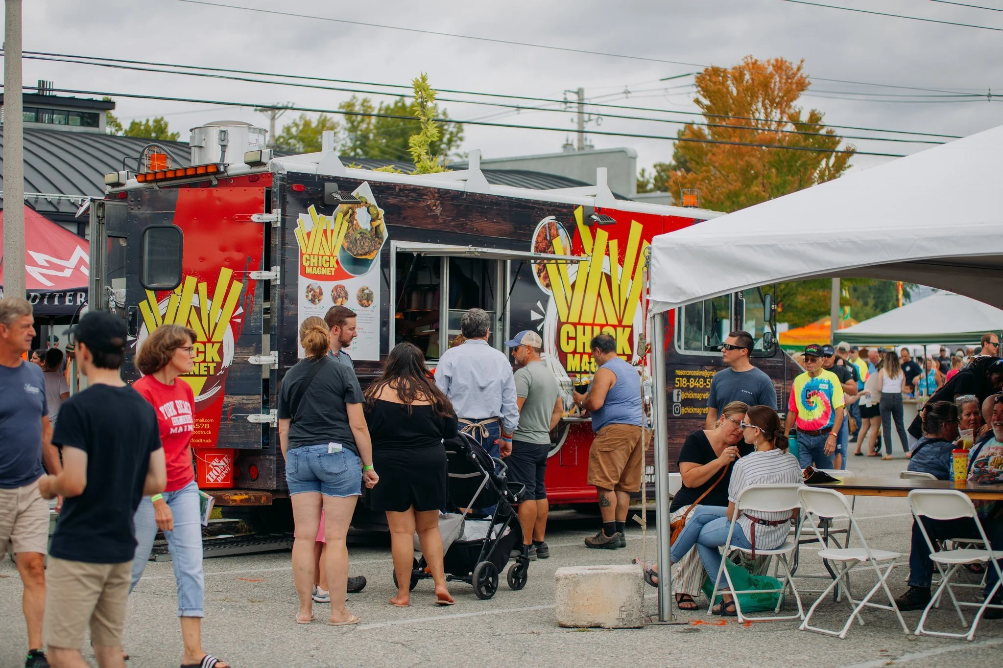 People standing in line and sitting at tables at a street food festival, with a food truck labeled Cheri Magnet and other tents in the background.