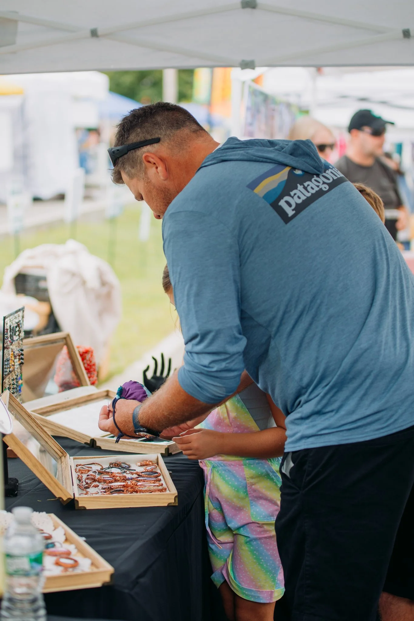 A man with short hair, wearing sunglasses on his head and a blue Patagonia hoodie, is looking at jewelry displayed on a table at an outdoor fair or market.