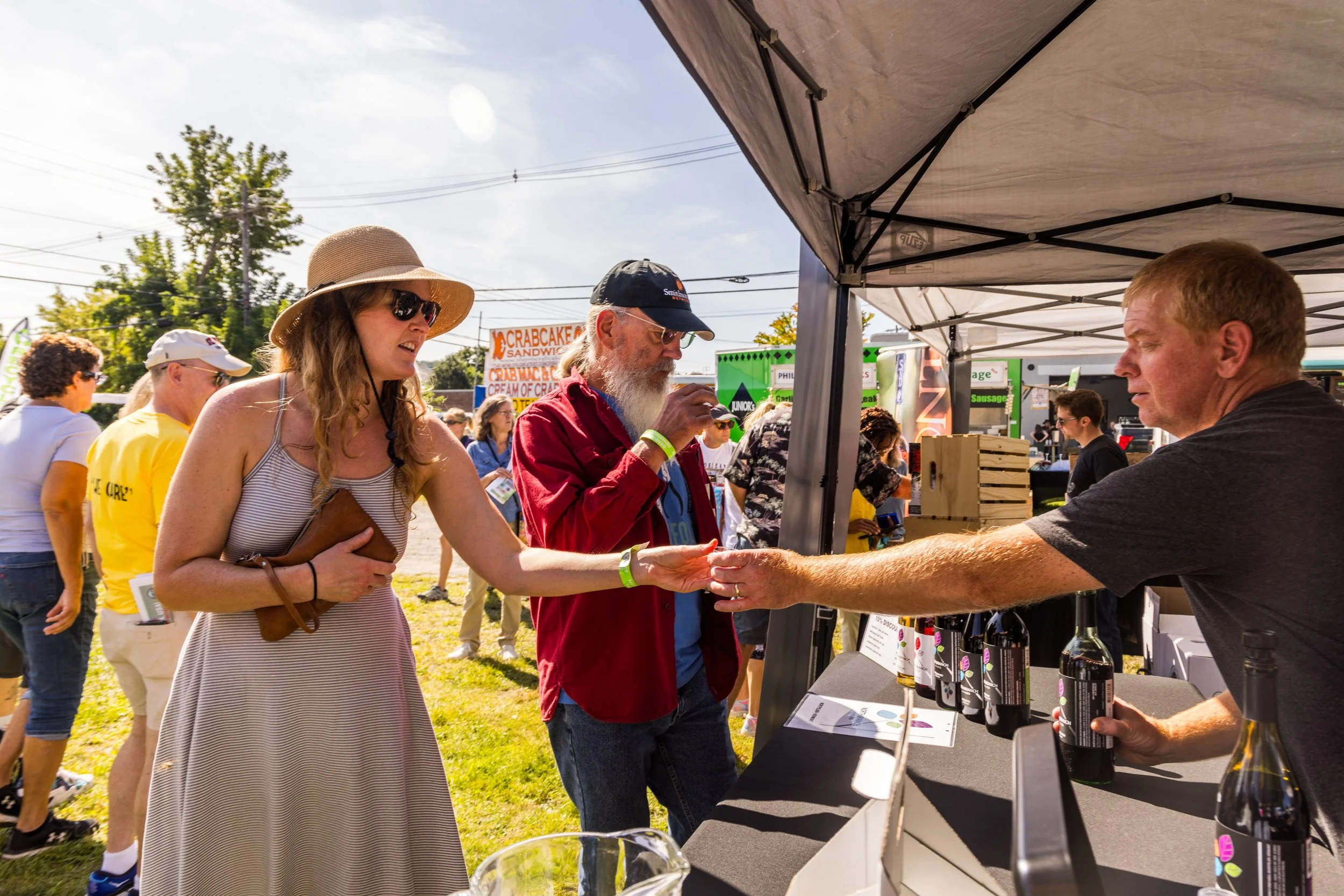 People at an outdoor food festival, a woman purchasing a bottle from a vendor at a booth, with others in line and food signs in the background.