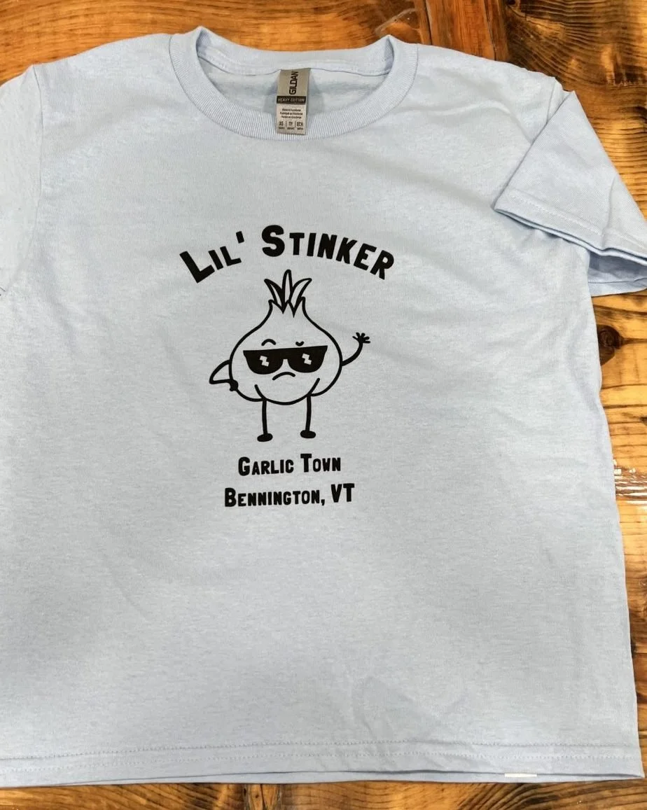 A light gray T-shirt with a cartoon garlic character wearing sunglasses and waving. The shirt has text that reads 'LIL' STINKER,' 'GARLIC TOWN,' and 'BENNINGTON, VT' on it.