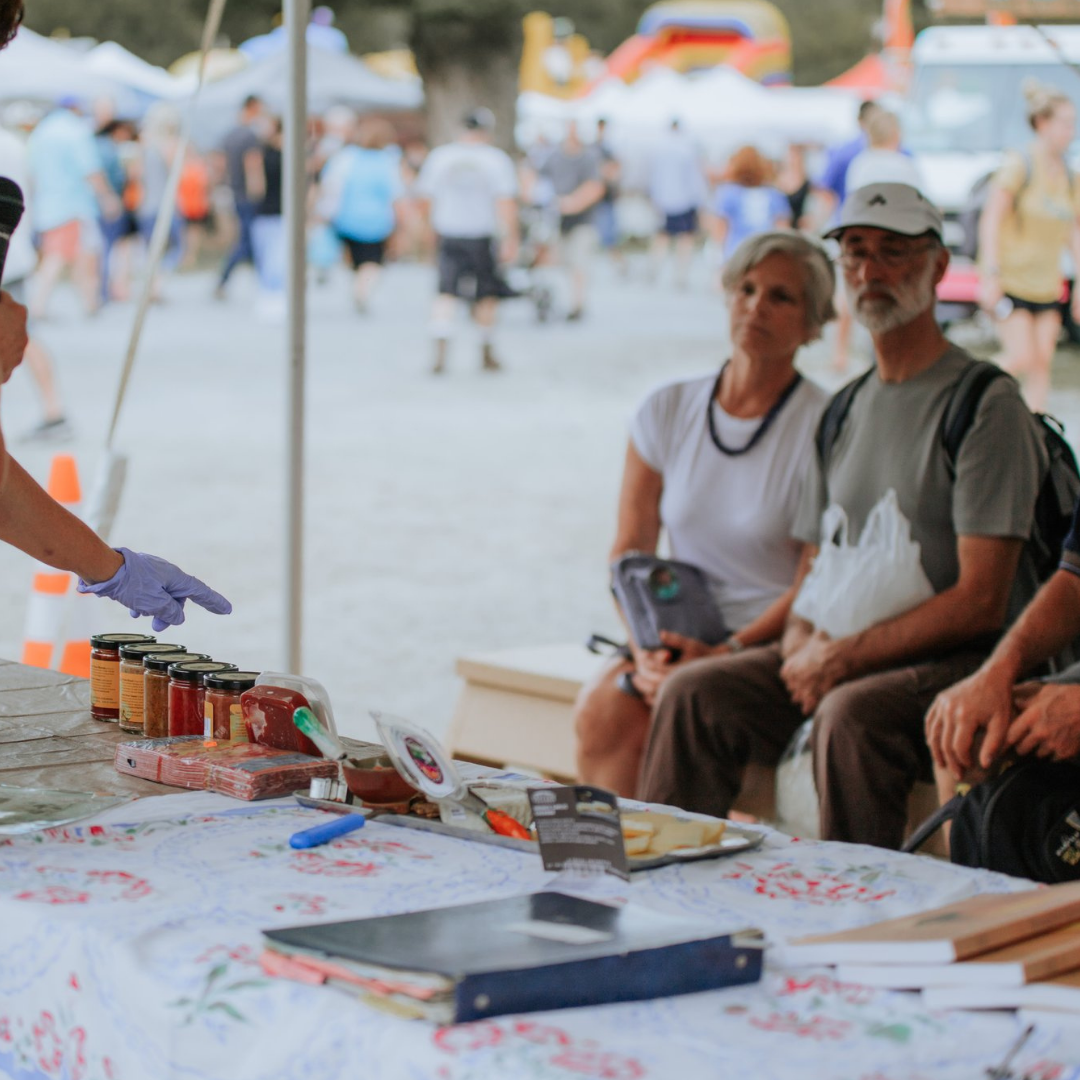 Two elderly people sitting at a table with jars and food items while at an outdoor market or festival. The background shows a busy scene with many people walking and tents.
