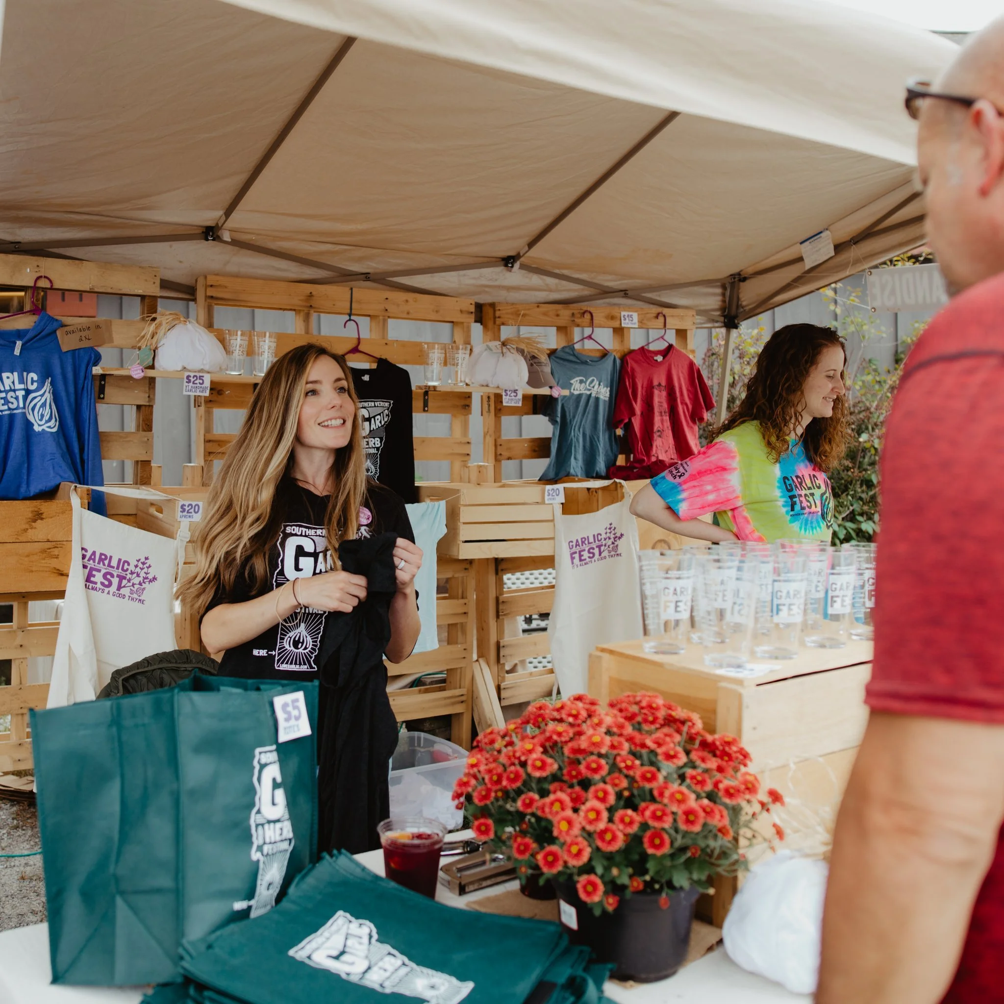 Two women selling merchandise at a Garlic Fest booth, with colorful shirts, tote bags, and glasses on display, and one woman smiling while holding a black shirt.