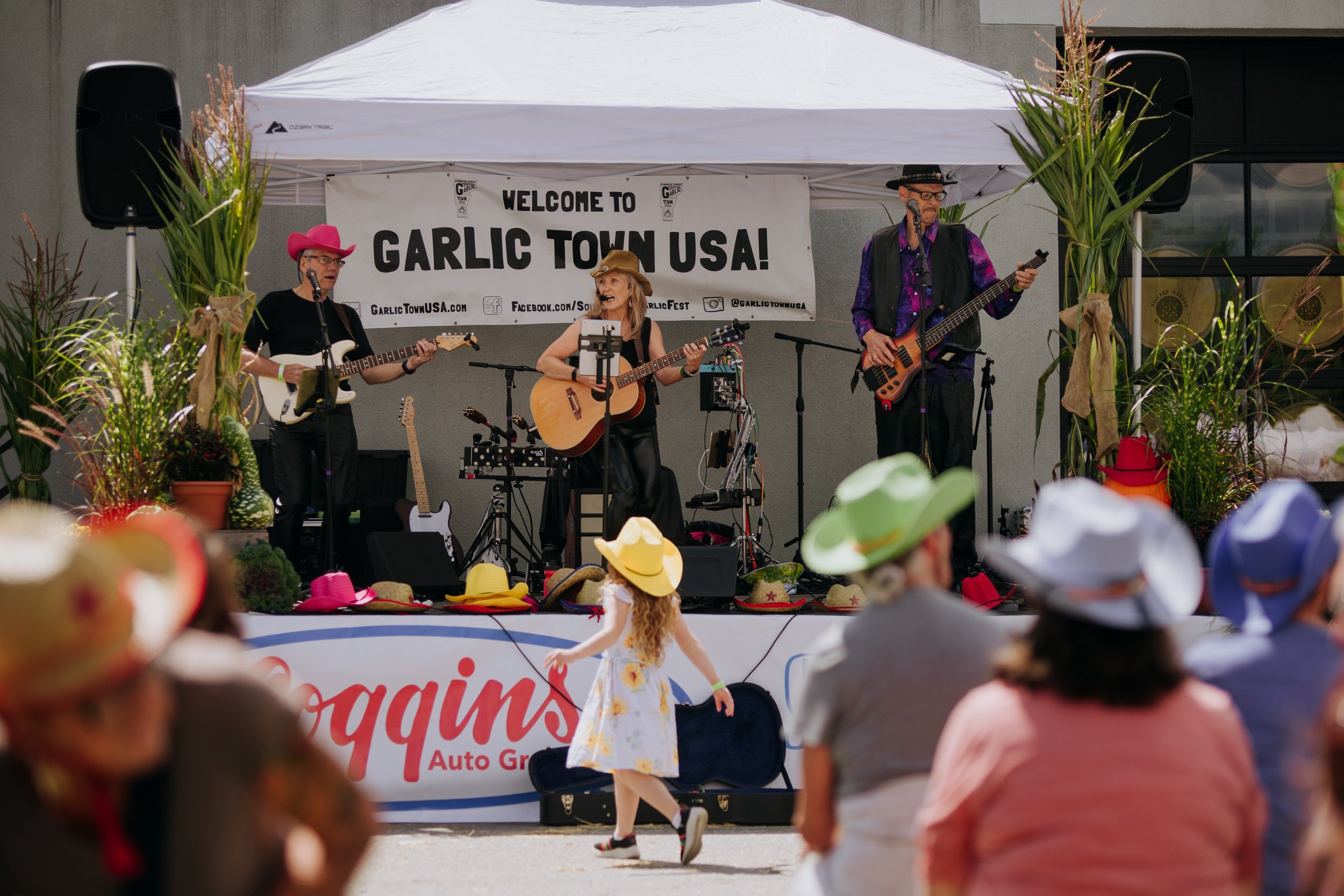 A band performing live on an outdoor stage at the Garlic Town USA event with a banner behind them. The stage is decorated with plants and hats, and there is an audience watching. A young girl in a yellow hat and dress is dancing in front of the stage.