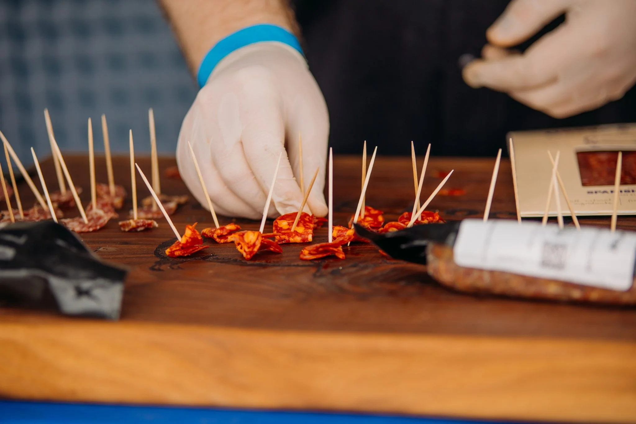 A person wearing white gloves is preparing small food appetizers with toothpicks on a wooden surface, with a packaged food item nearby.
