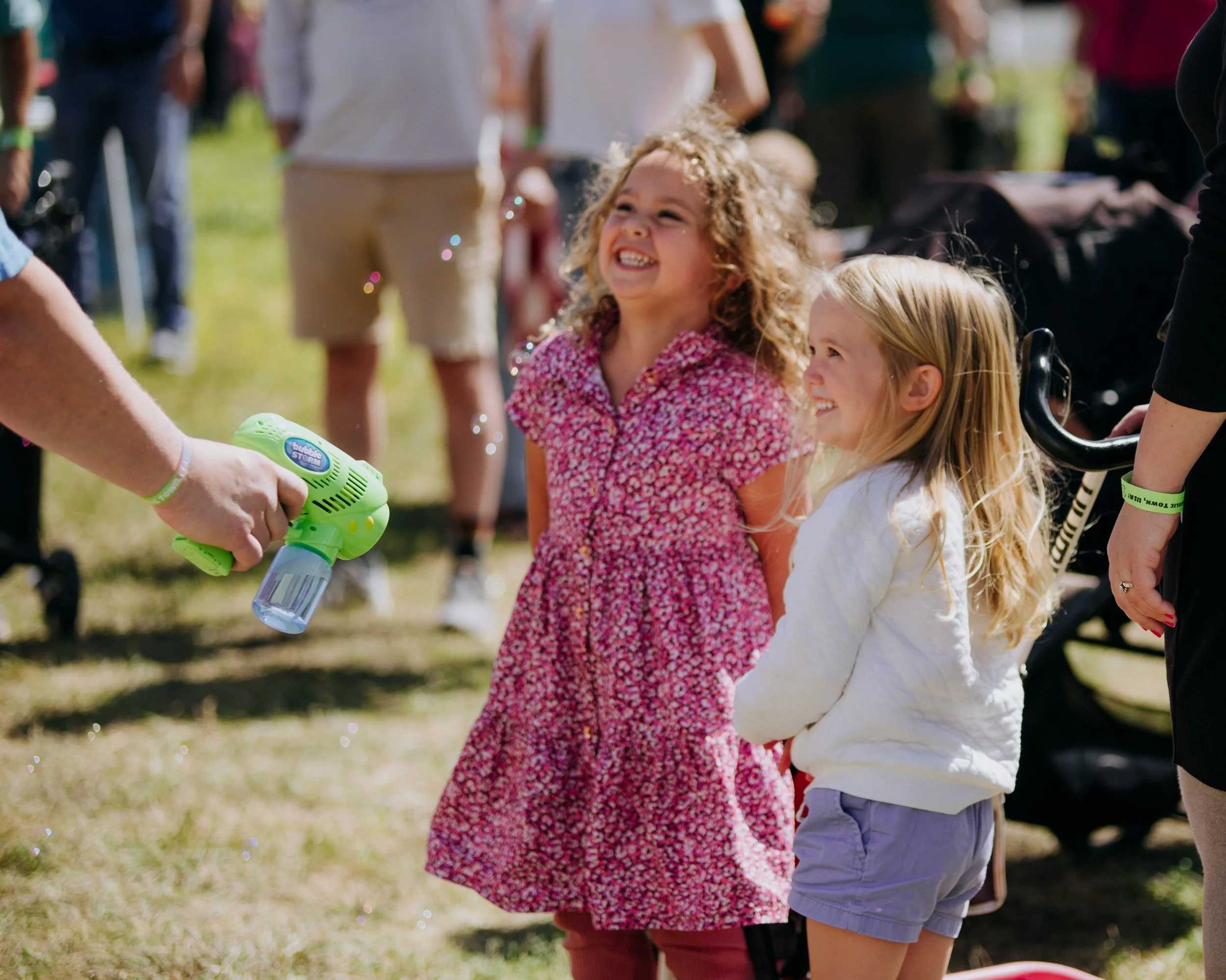 Two young girls smiling and laughing as one is being sprayed with bubbles from a bubble machine at an outdoor gathering.