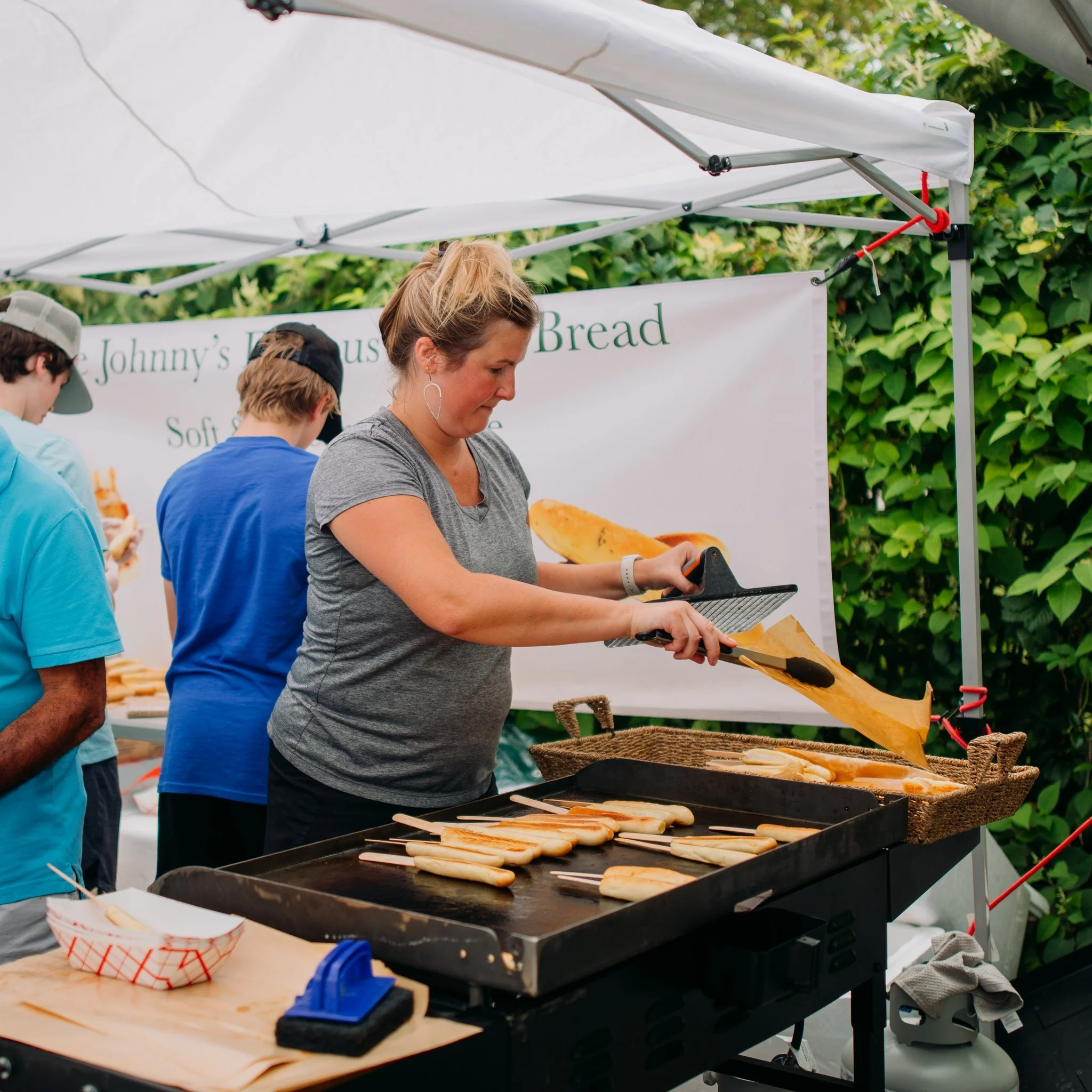 A woman is cooking hot dogs on a flat-top grill at an outdoor food stall, with a banner in the background advertising bread and other foods, and there are other people serving and browsing the stall.