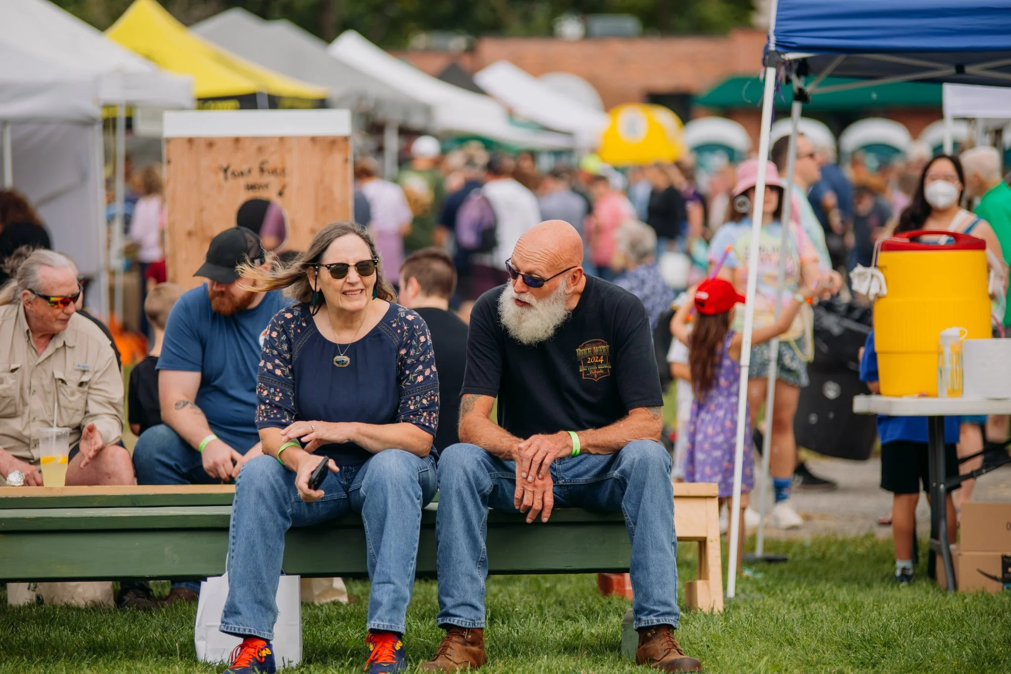 Two people sitting on a bench at an outdoor event, talking and smiling, with a crowd of people and vendor tents in the background.