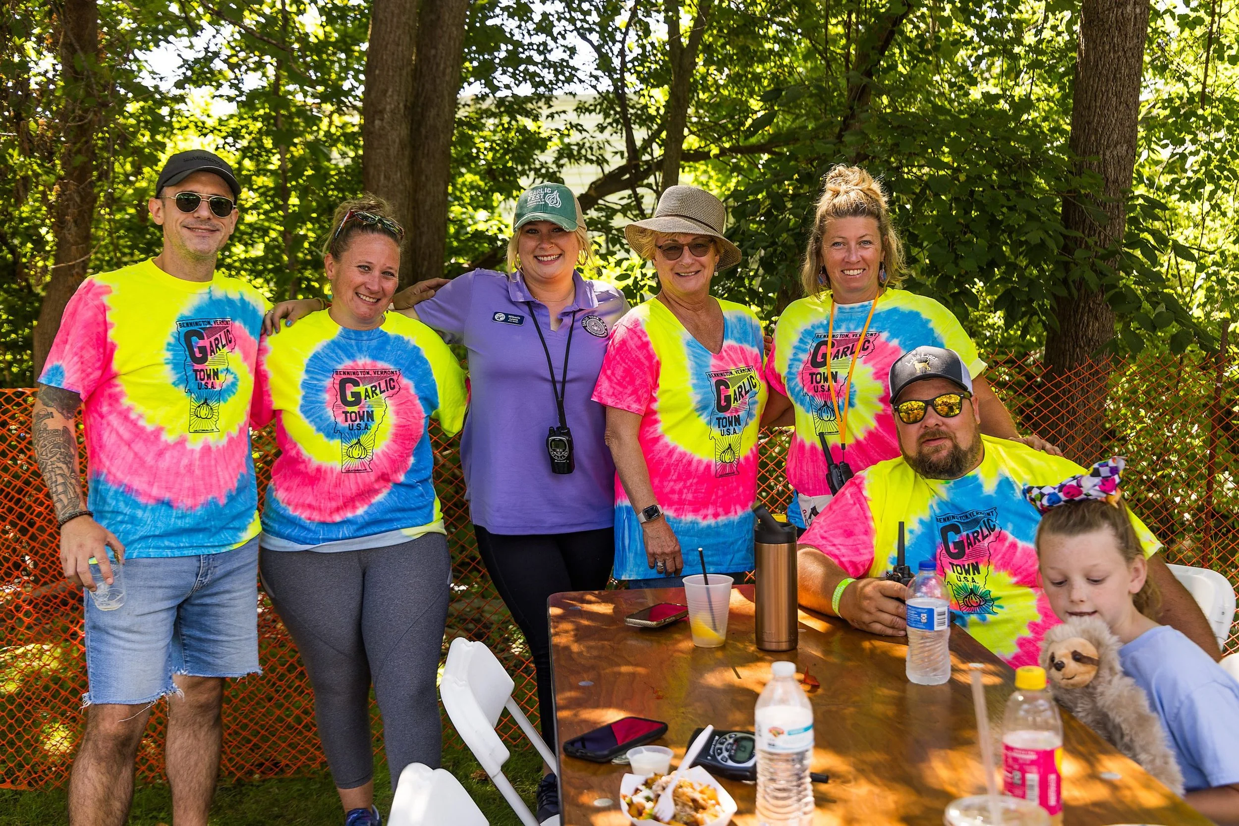 Group of people outdoors, some wearing colorful tie-dye shirts with 'Garlic Town' logo, standing and sitting around a wooden table with food and drinks, surrounded by trees and orange safety fencing.