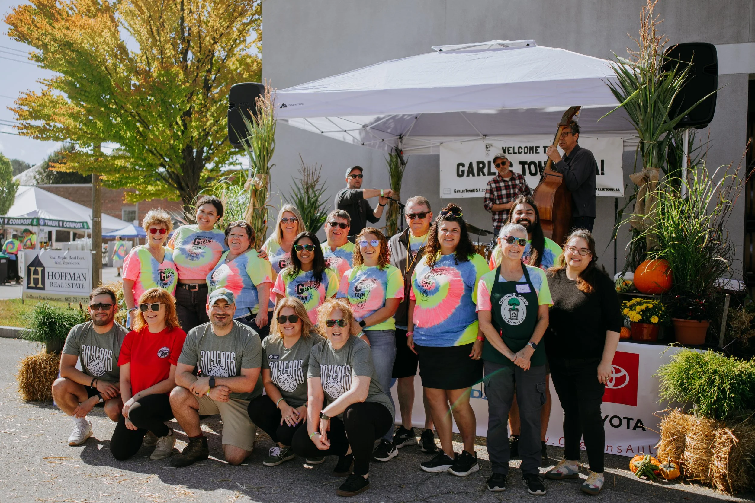 Group of people smiling at a fall festival, some wearing colorful tie-dye shirts, standing in front of a decorated stage with a band playing, pumpkins, hay bales, and fall plants, with tents and trees in the background.