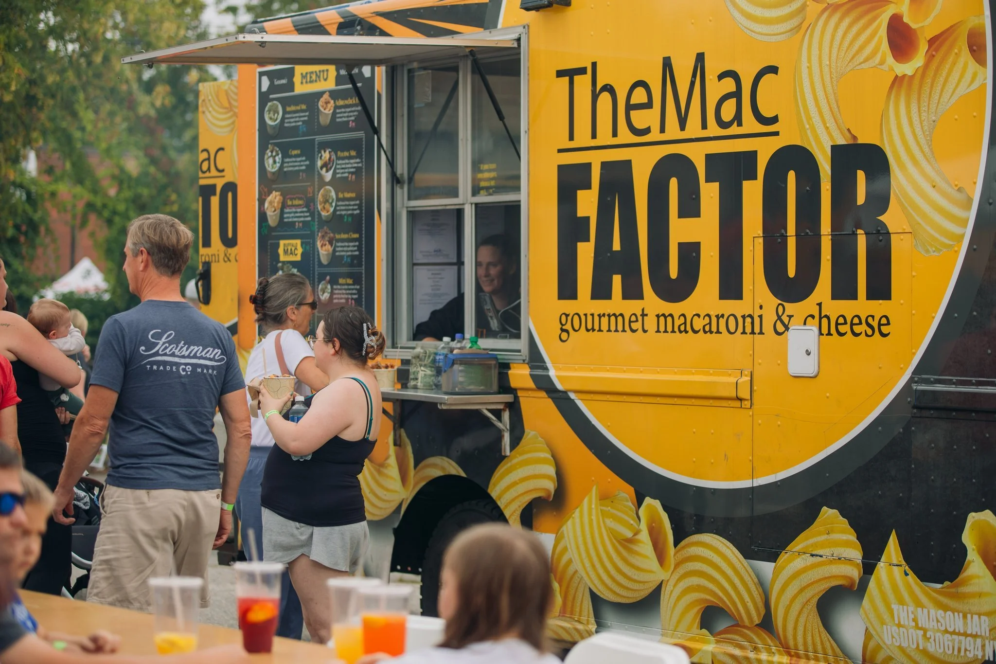 A yellow food truck named The Mac Factor, serving gourmet macaroni and cheese. People are standing in line and interacting with staff. Several drinks are on a table in the foreground.