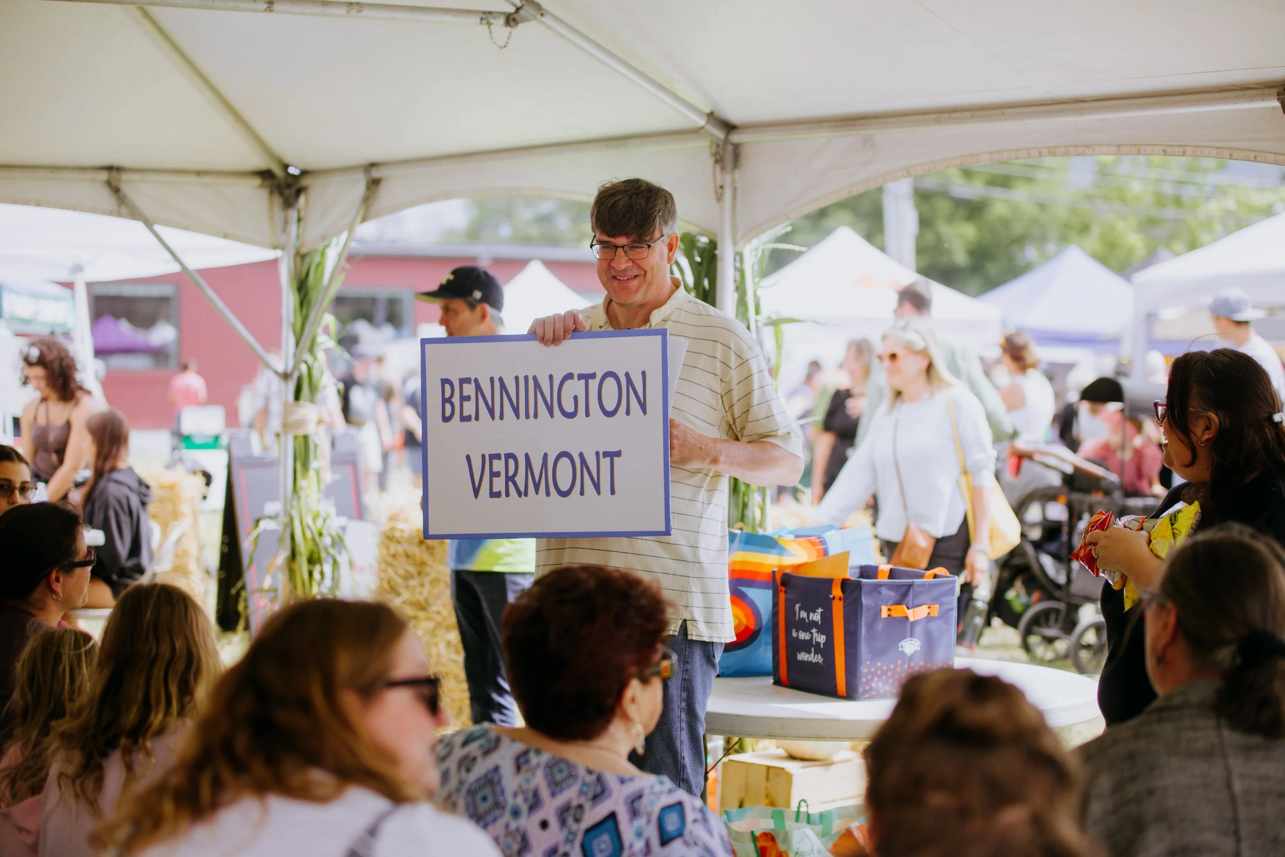 A man holding a sign that reads "Bennington Vermont" at an outdoor market or fair, with people sitting at tables and walking around in the background under tent canopies.