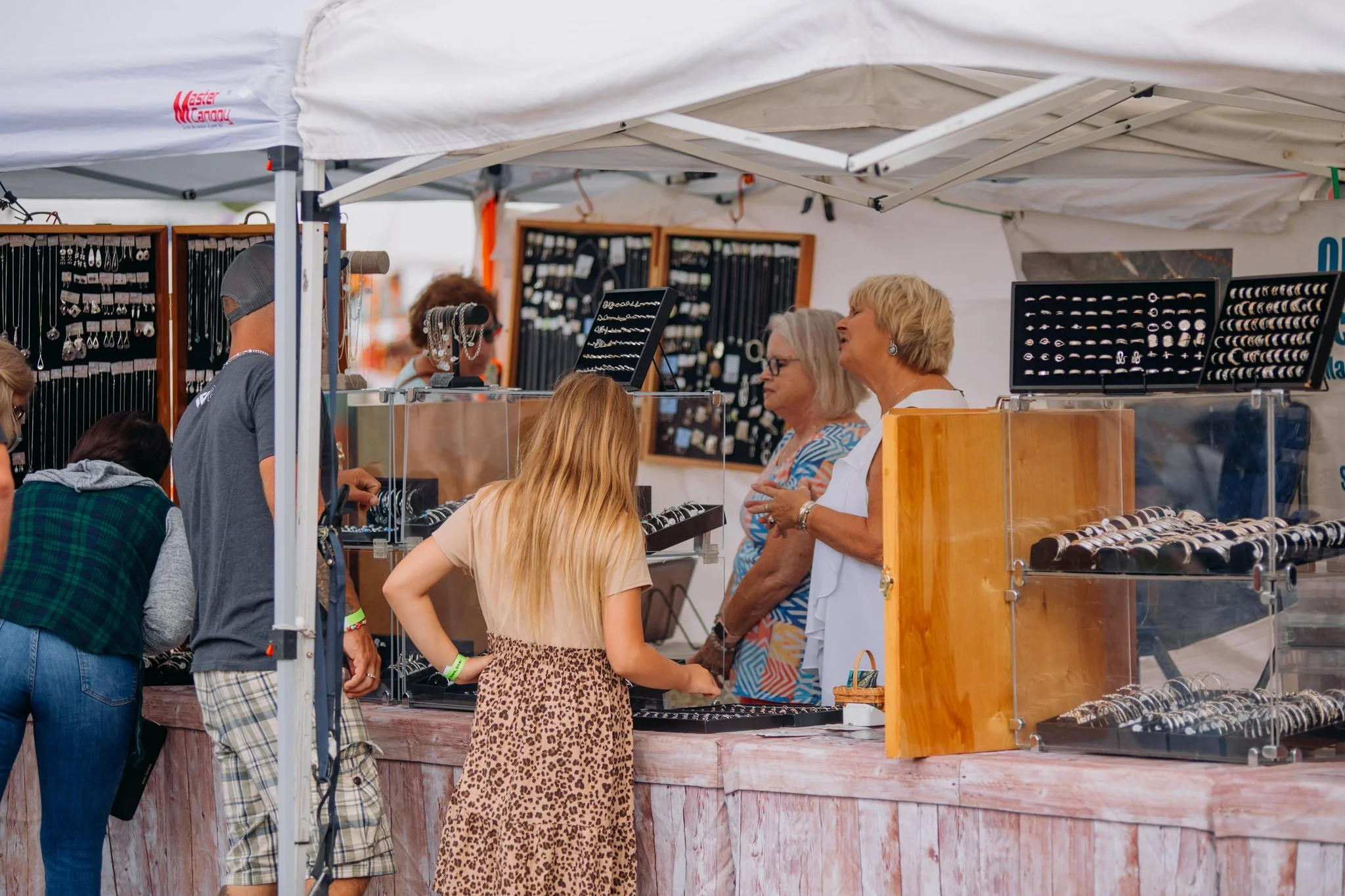 People shopping for jewelry at an outdoor market stall with display cases and jewelry hanging on boards.