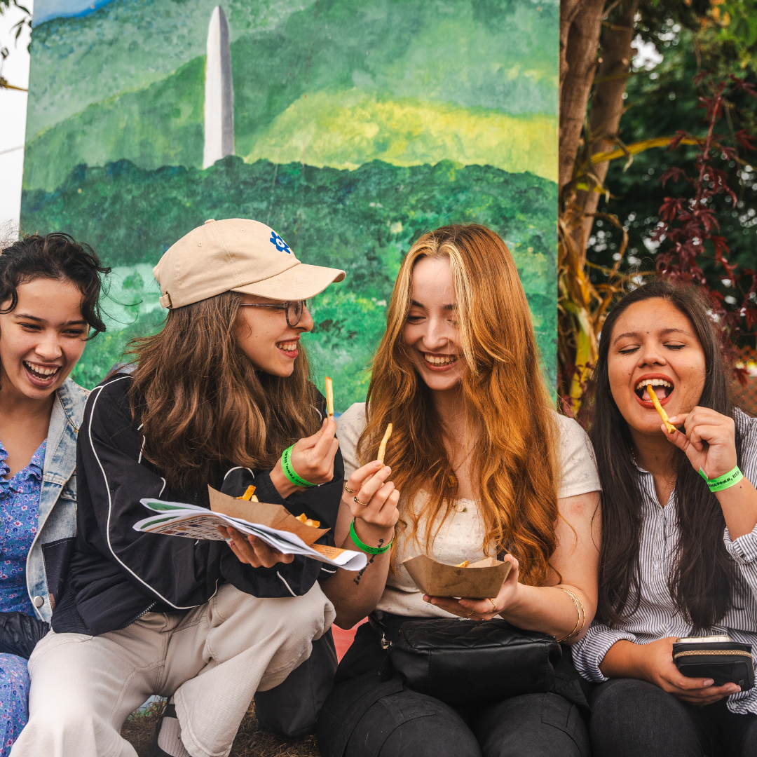 Four young women laughing and eating French fries outdoors in front of a painted mural of the Washington Monument and green landscape.