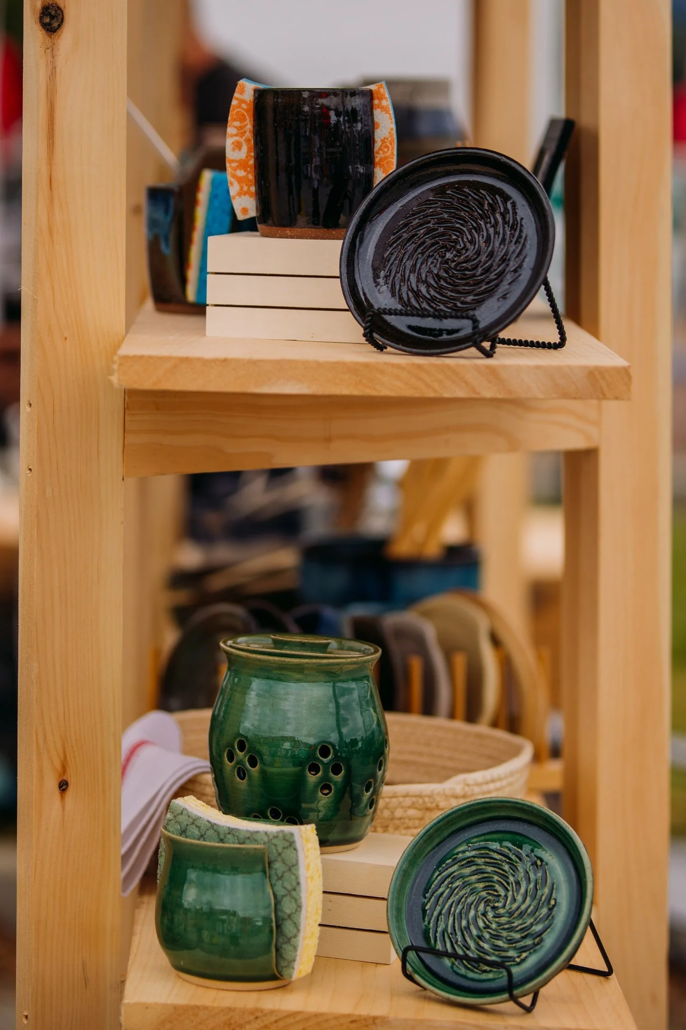 Wooden shelf displaying various ceramic pottery items, including green vases and black and green plates with fingerprint designs, some arranged on small white stands.