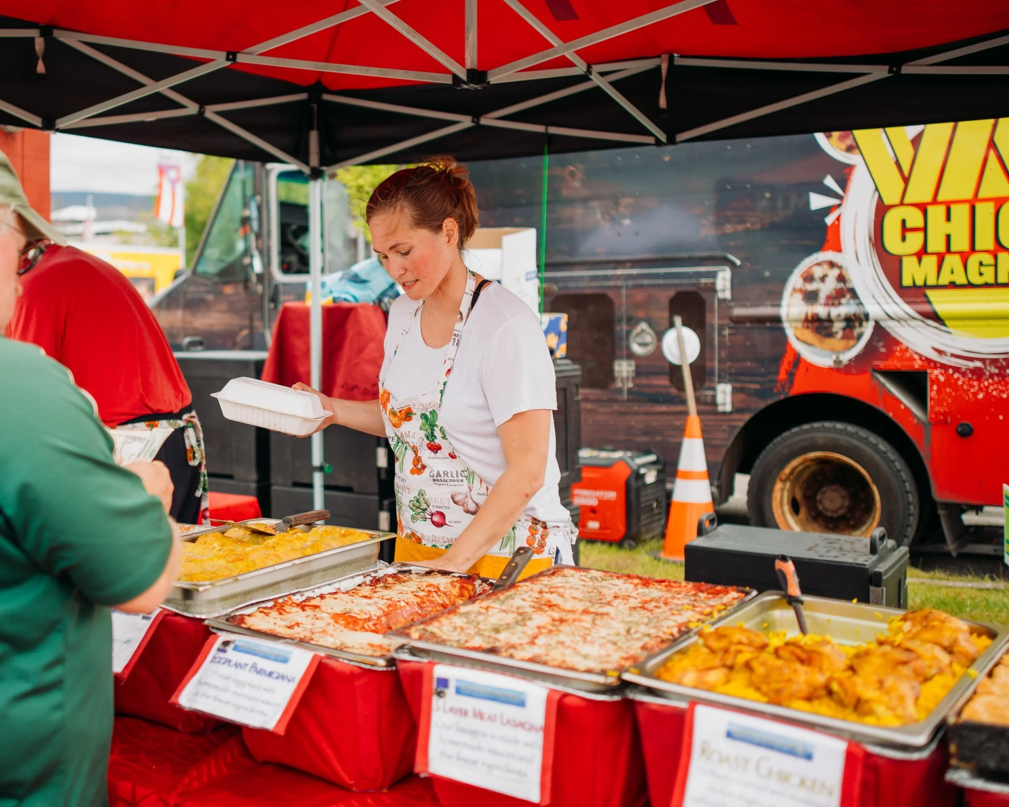 Food vendor at a food truck festival serving dishes at a food stall with trays of cooked food, under a red canopy.