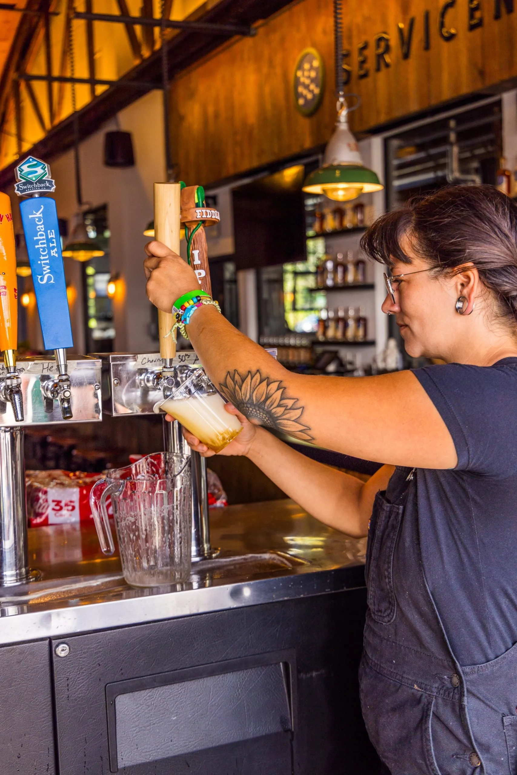 A woman with glasses and a tattoo on her arm is pouring a beer from a tap at a bar, with multiple beer taps in front of her and a wooden wall in the background.