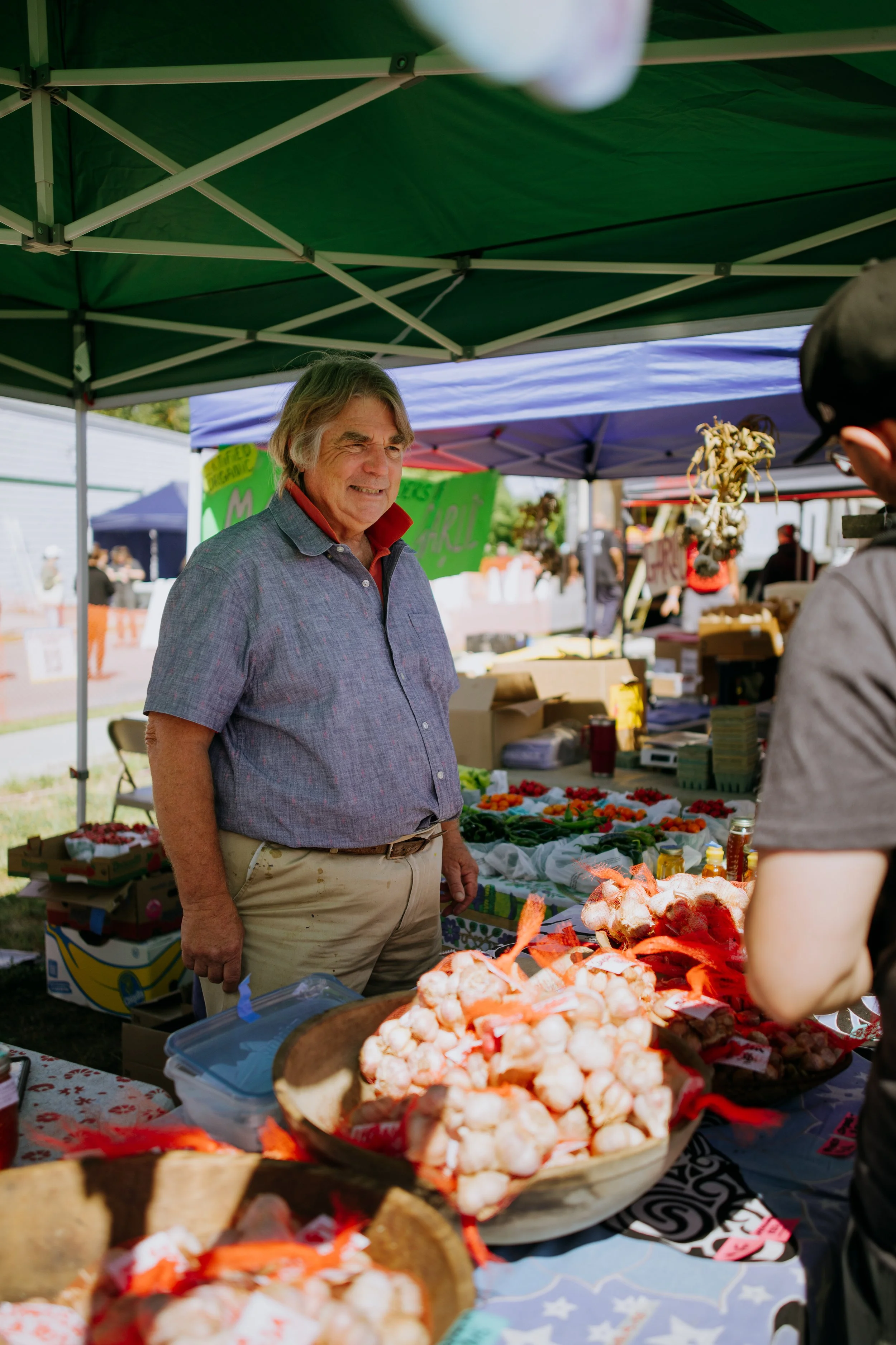 An elderly man smiling at a farmer's market stall selling garlic and vegetables under a green canopy.