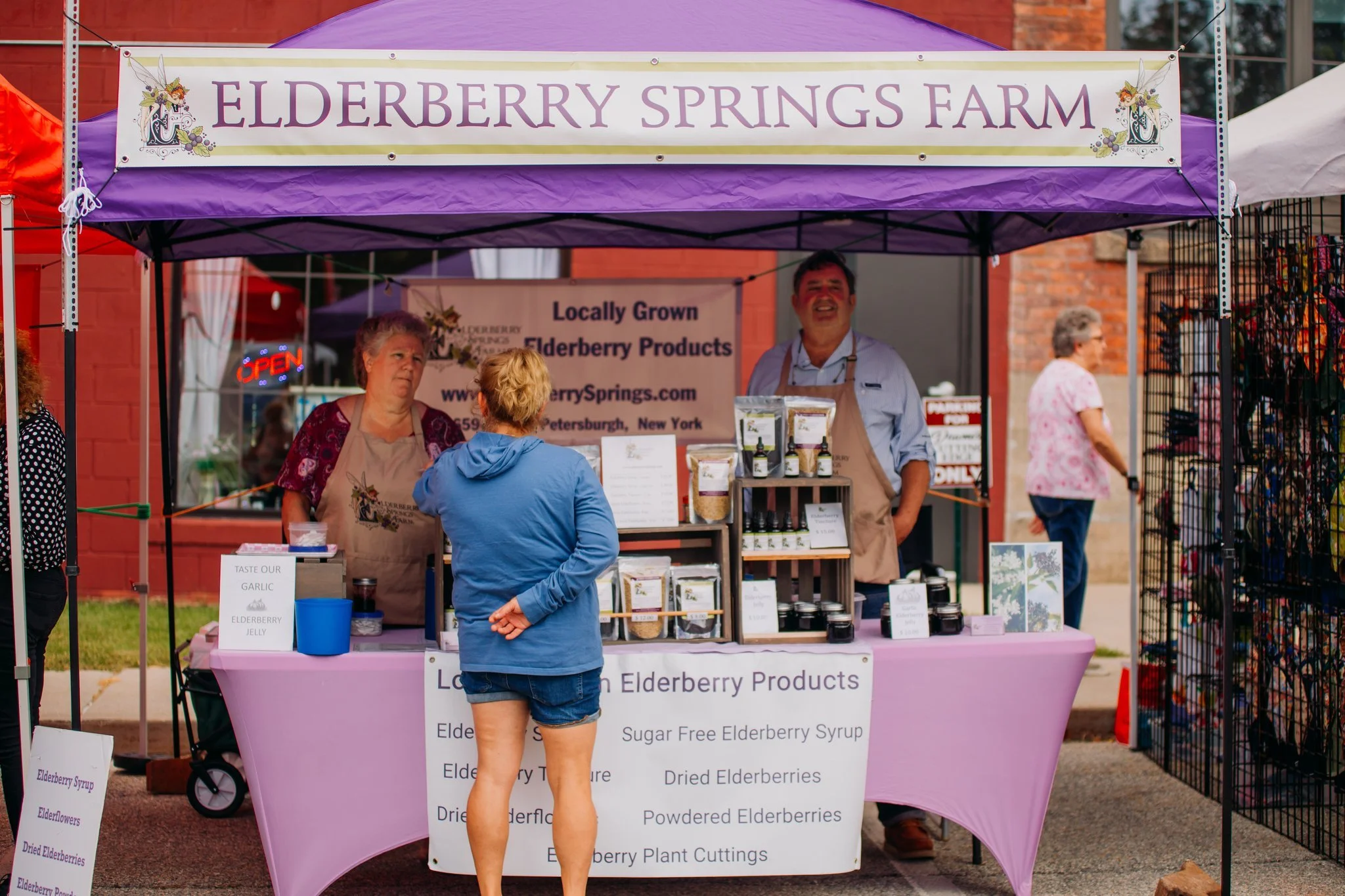 A market stall with a purple canopy labeled Elderberry Springs Farm selling elderberry products. Two vendors, a woman and a man, stand behind the stall, which displays jars and bags of elderberry items. A customer in a blue hoodie is talking to the v