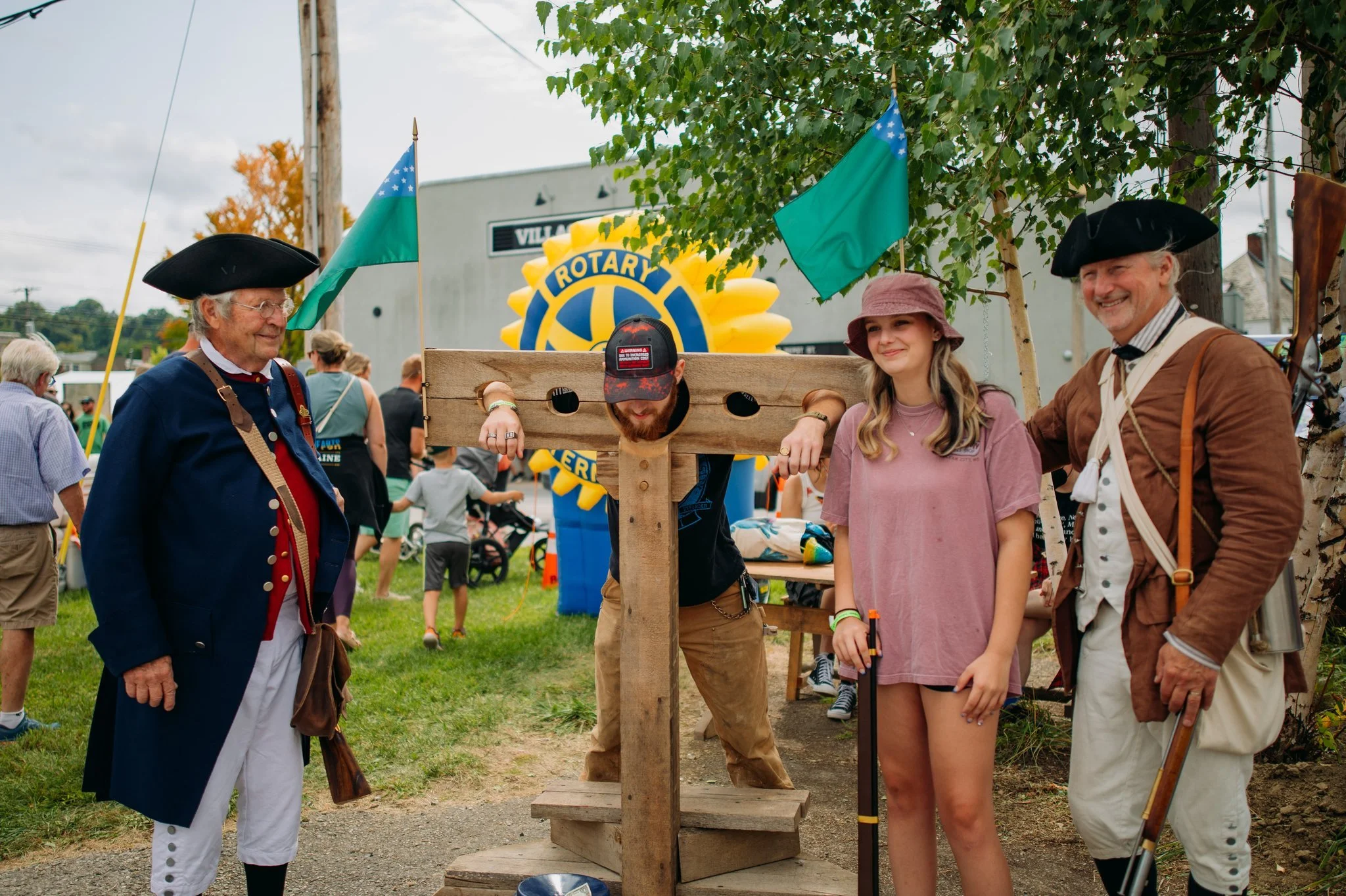 People dressed in historical costumes participating in a reenactment or event, with a person on a wooden pillory and two flags, outdoors with a crowd and a Rotary sign in the background.