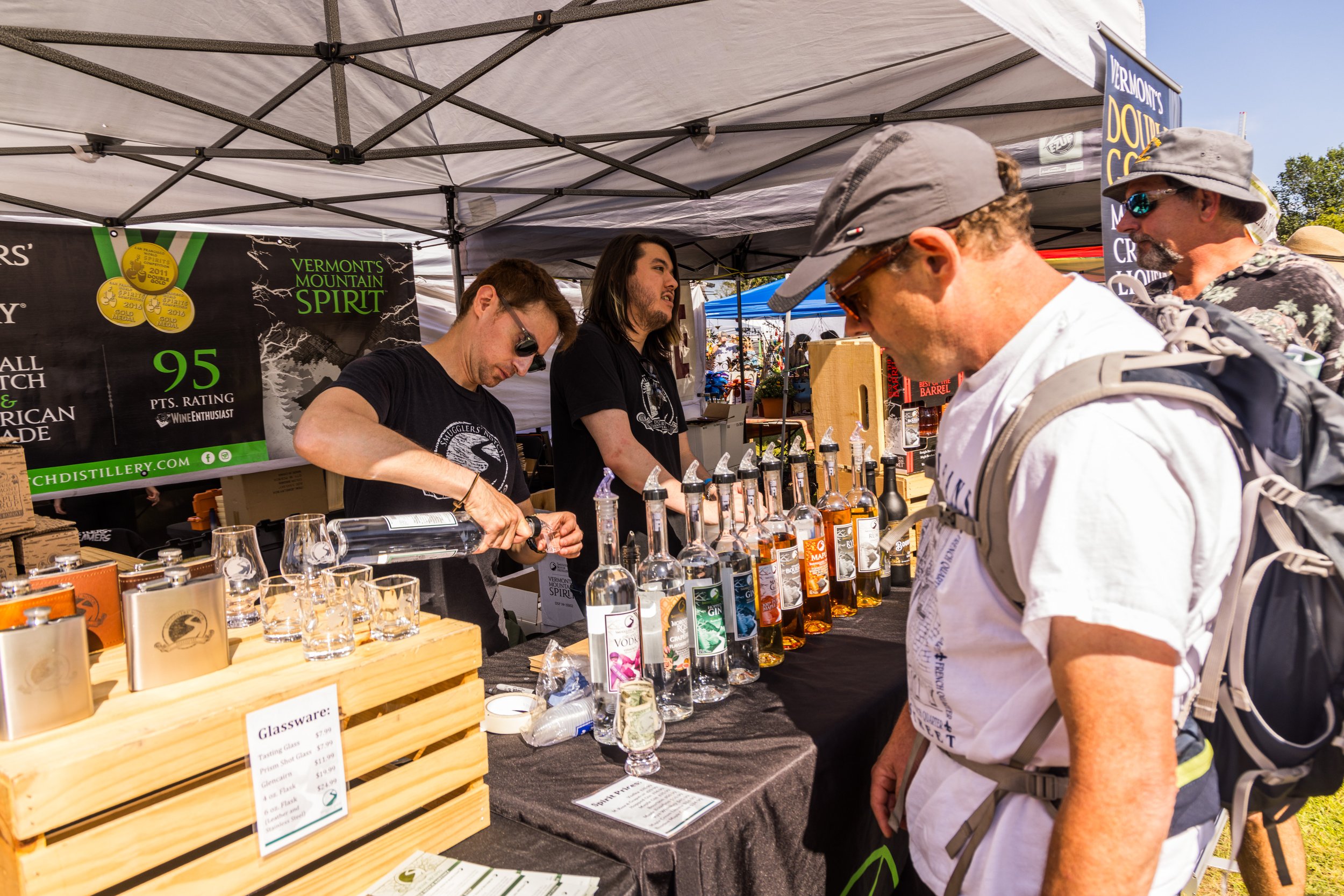 Two men and two women at a tasting booth under a white canopy tent, sampling and selling bottles of spirits, with glasses and a black banner displaying awards and ratings in the background.