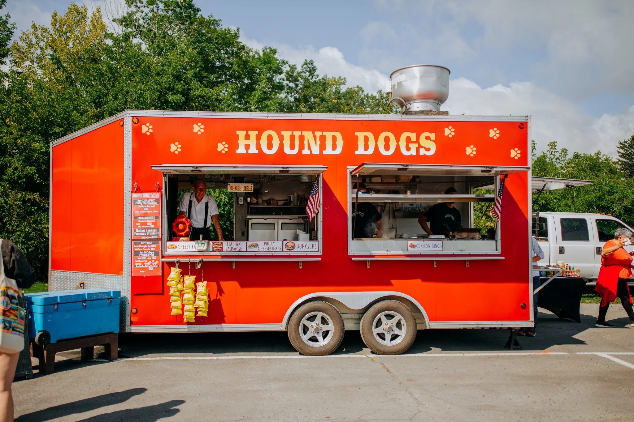 Red food truck with 'HOUND DOGS' sign and paw print decorations, serving hot dogs and other items at an outdoor event with people nearby and trees in the background.