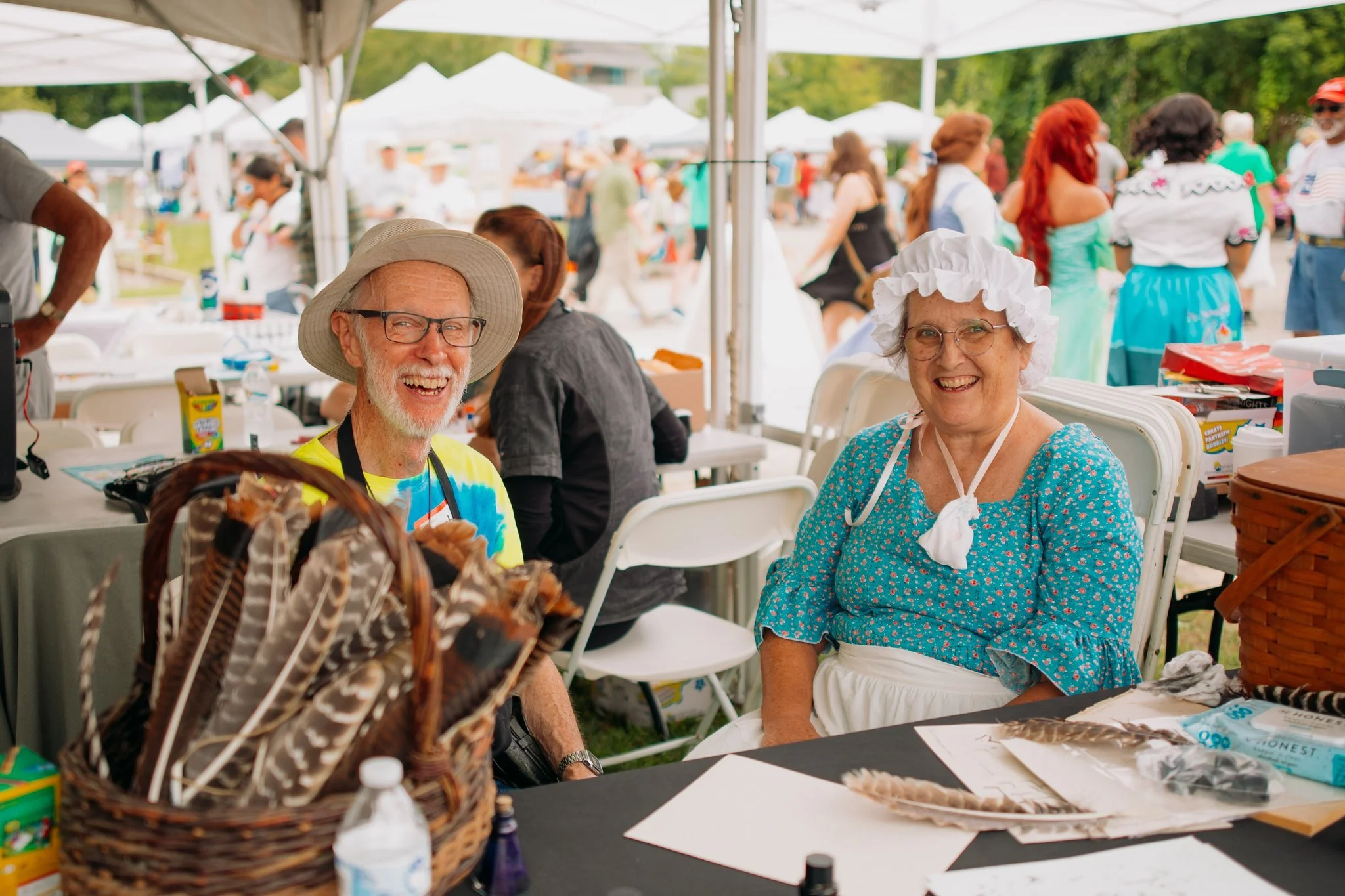 Smiling elderly man and woman dressed in vintage attire sitting at a table under a tent at an outdoor event, surrounded by other people and booths.