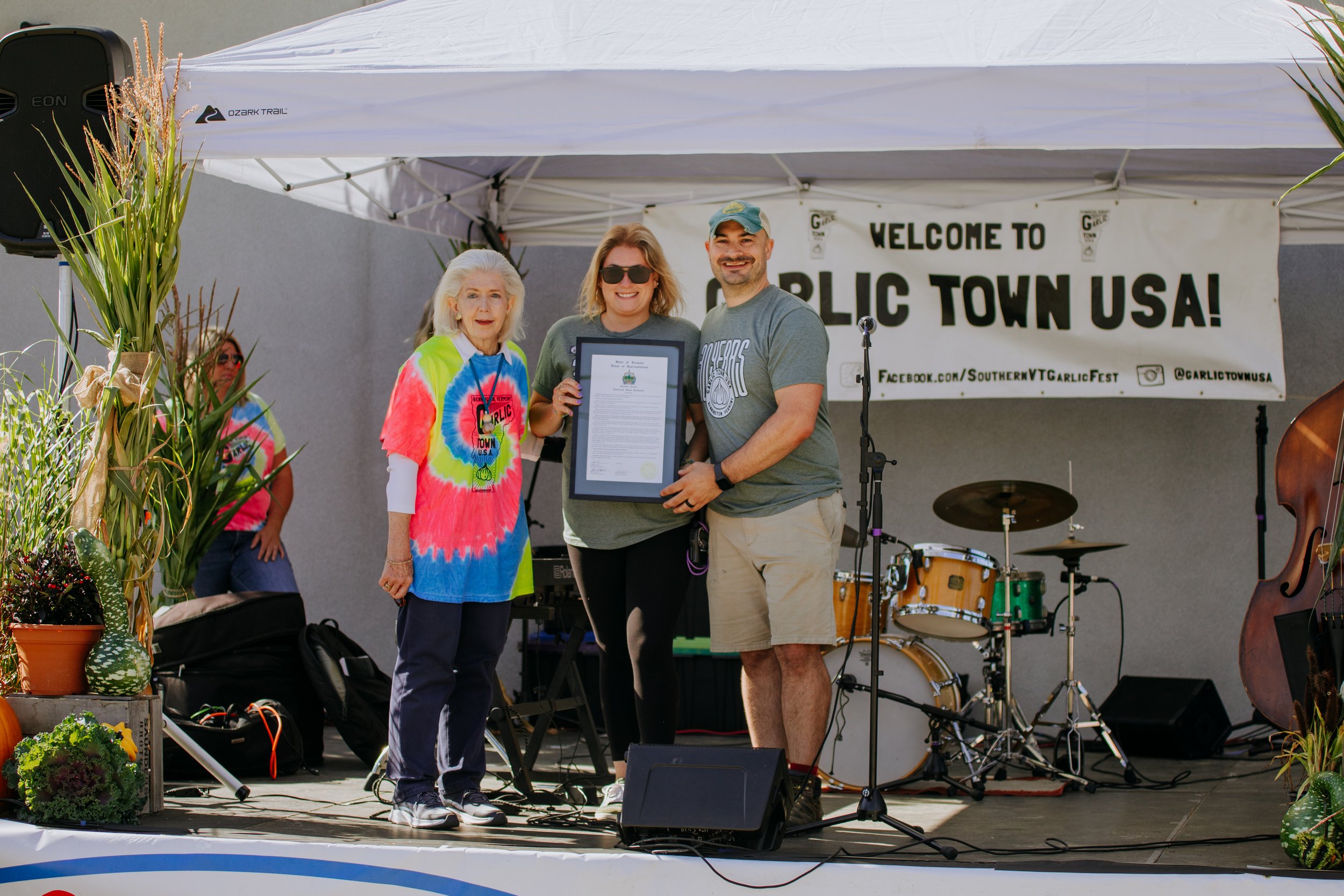 Three people on a stage at a city event, with two women and one man, holding a framed document and smiling. The backdrop has a banner welcoming to Garlic Town USA, with musical instruments and plants visible on stage.