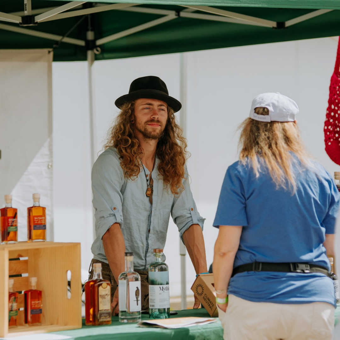A man with long curly hair, a beard, wearing a black hat and a light blue shirt, stands at an outdoor market stall with bottles of alcohol displayed on the table. A woman with blond hair, wearing a white cap and a blue shirt, is facing him. The stall