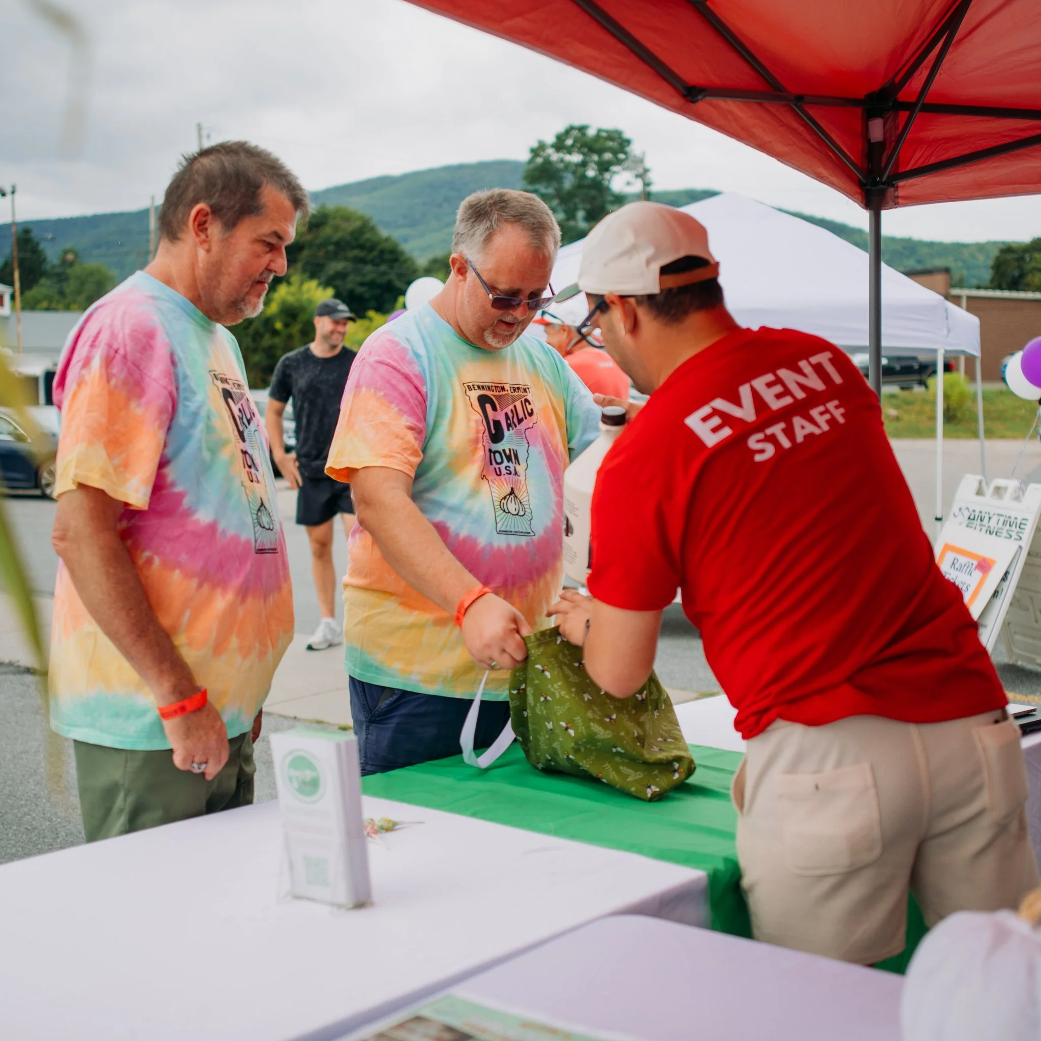 At an outdoor event, a man in a red event staff shirt hands a green gift bag to a man in a tie-dye t-shirt. Two other men in colorful tie-dye shirts watch. A man in the background looks on, with tents and trees in the distance.