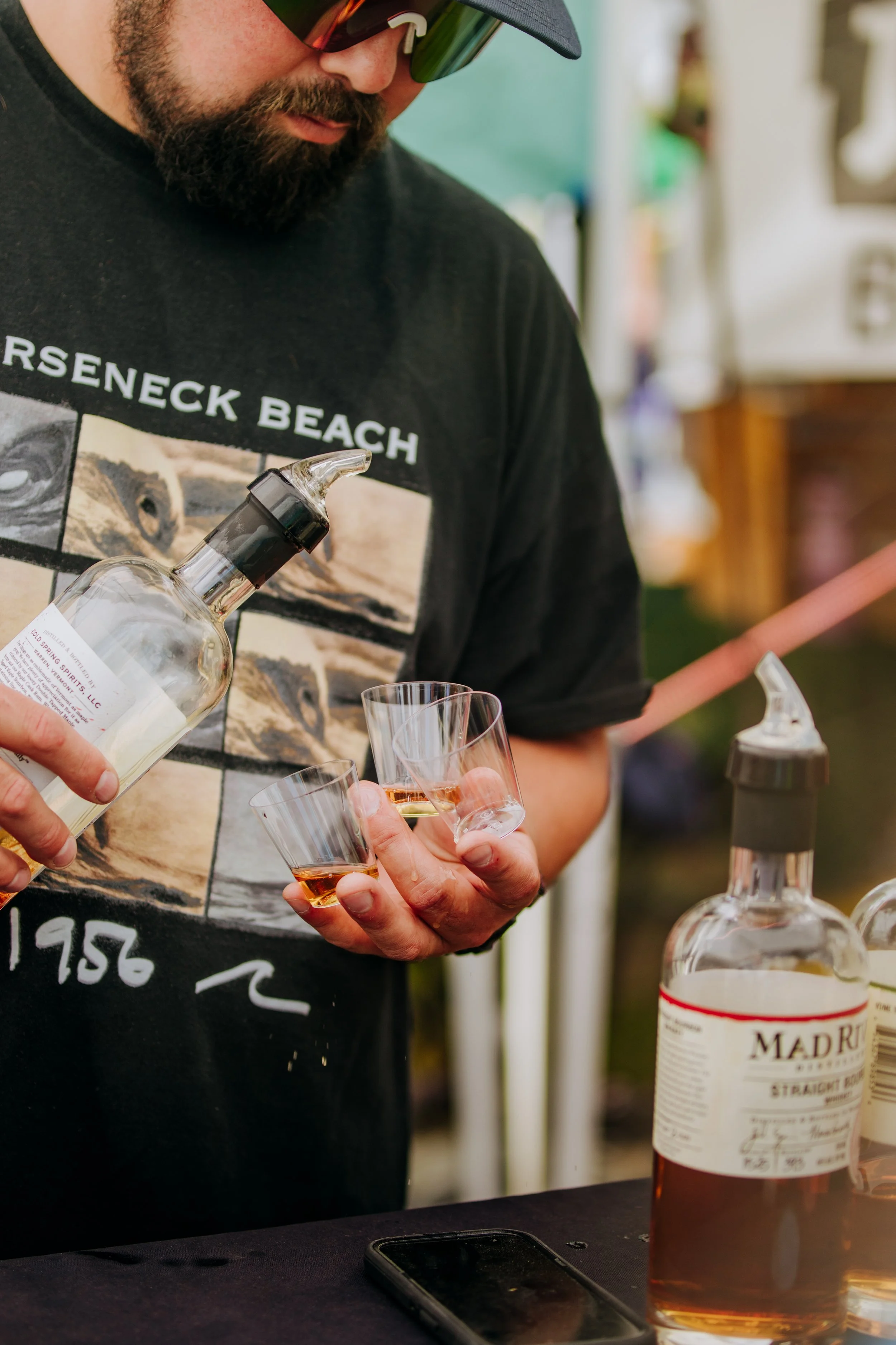 A man in sunglasses and a black t-shirt pouring whiskey into small glasses at a bar or outdoor event.