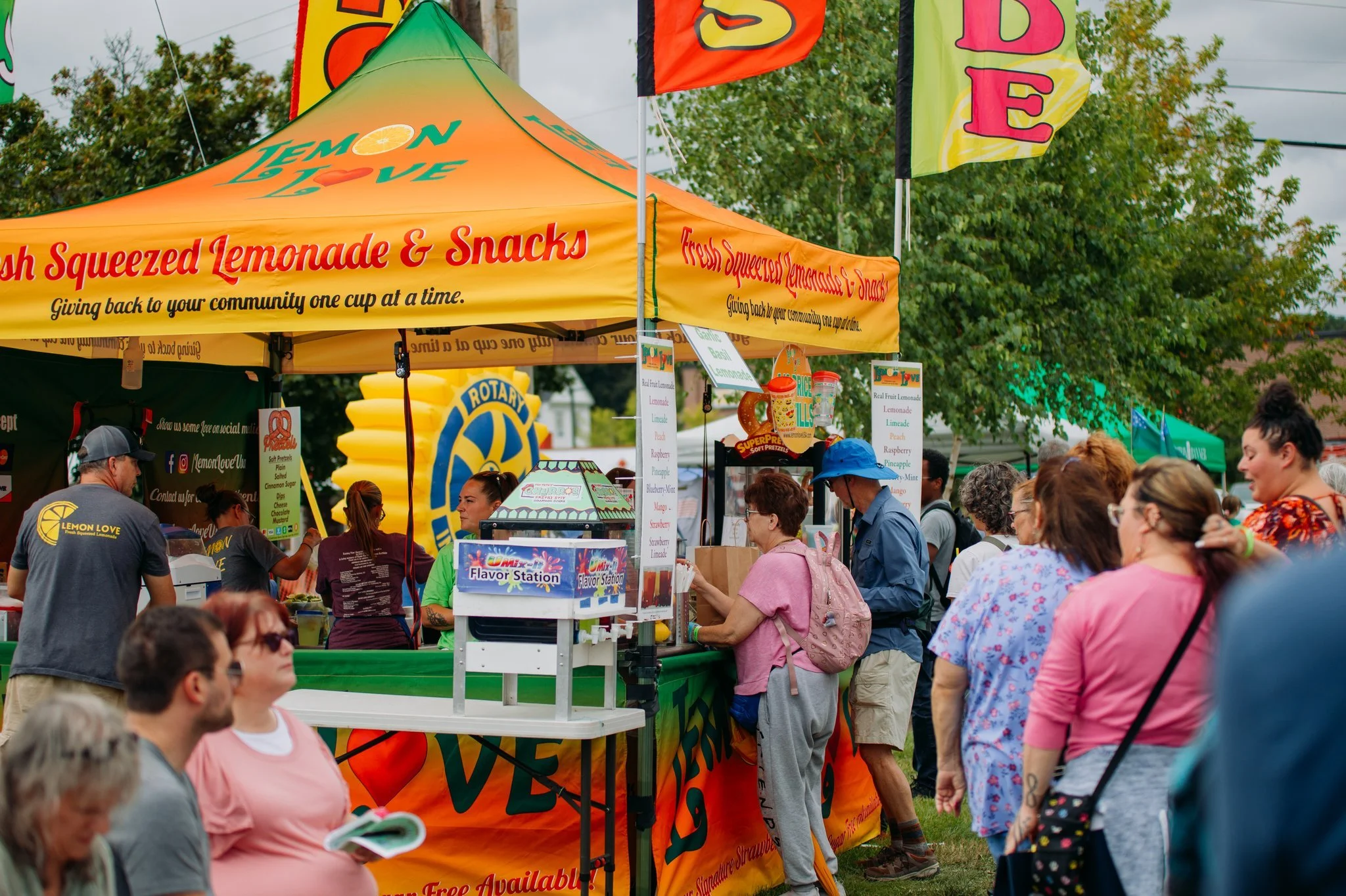 A crowded outdoor fair with a Lemonade stand and many people gathering around