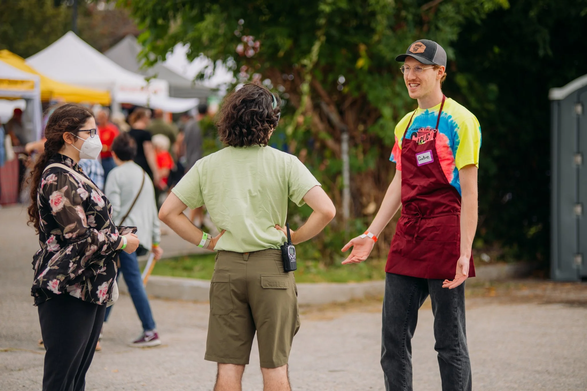 Three people having a conversation outdoors at an event, with tents in the background. One person is wearing a mask and glasses, another in a green t-shirt with a radio clipped to shorts, and the third person is wearing a colorful tie-dye shirt, a re