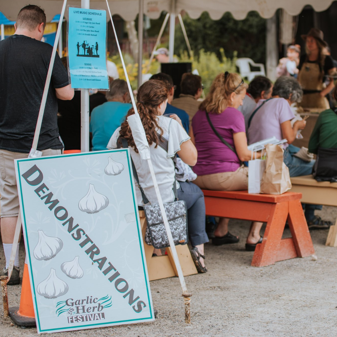 A crowd seated on benches at the Garlic Herb Festival, with a sign indicating demarcation for the demonstration area and a music schedule visible in the background.