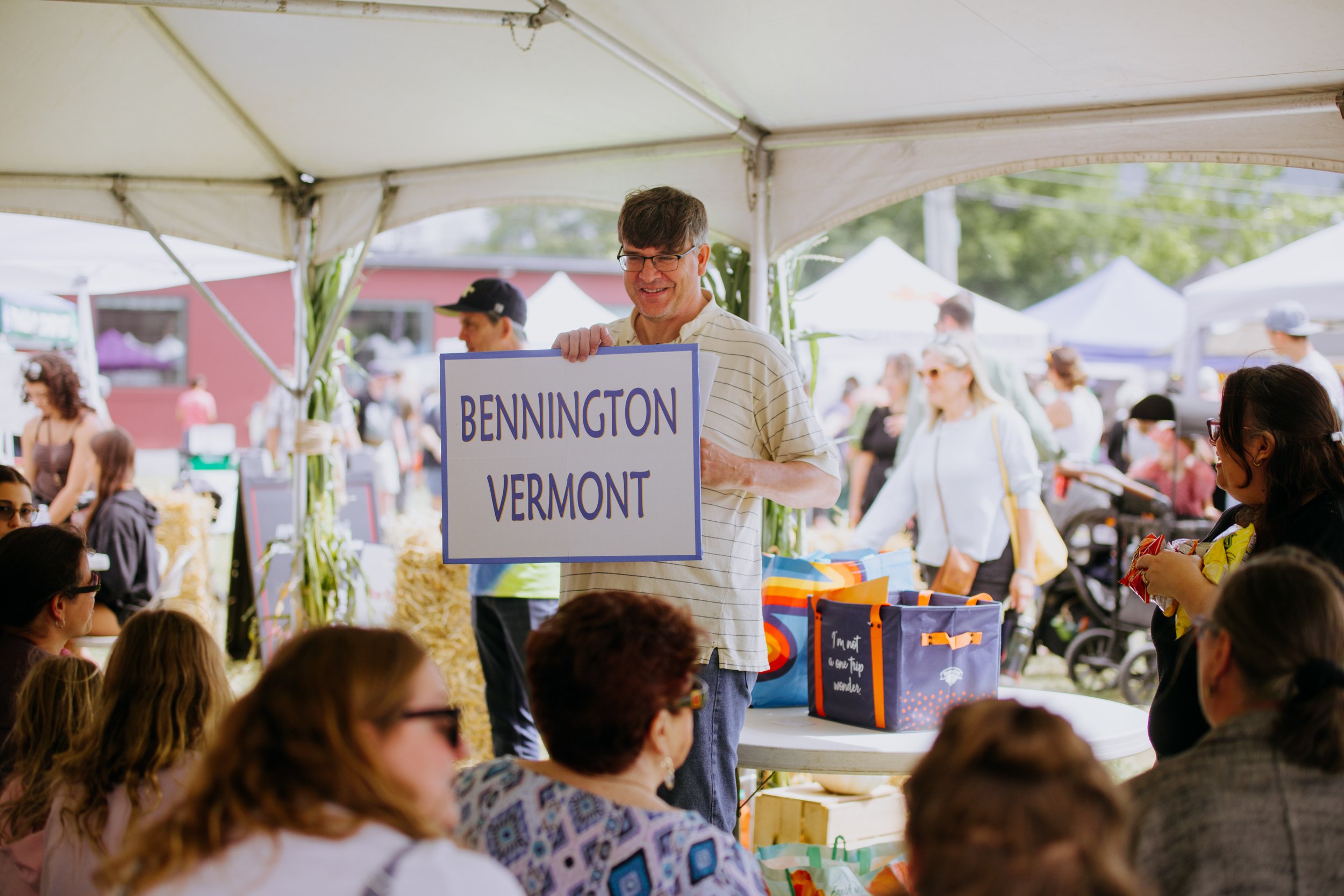 A man holding a sign that says Bennington Vermont at an outdoor event or festival under a tent, with people in the background.