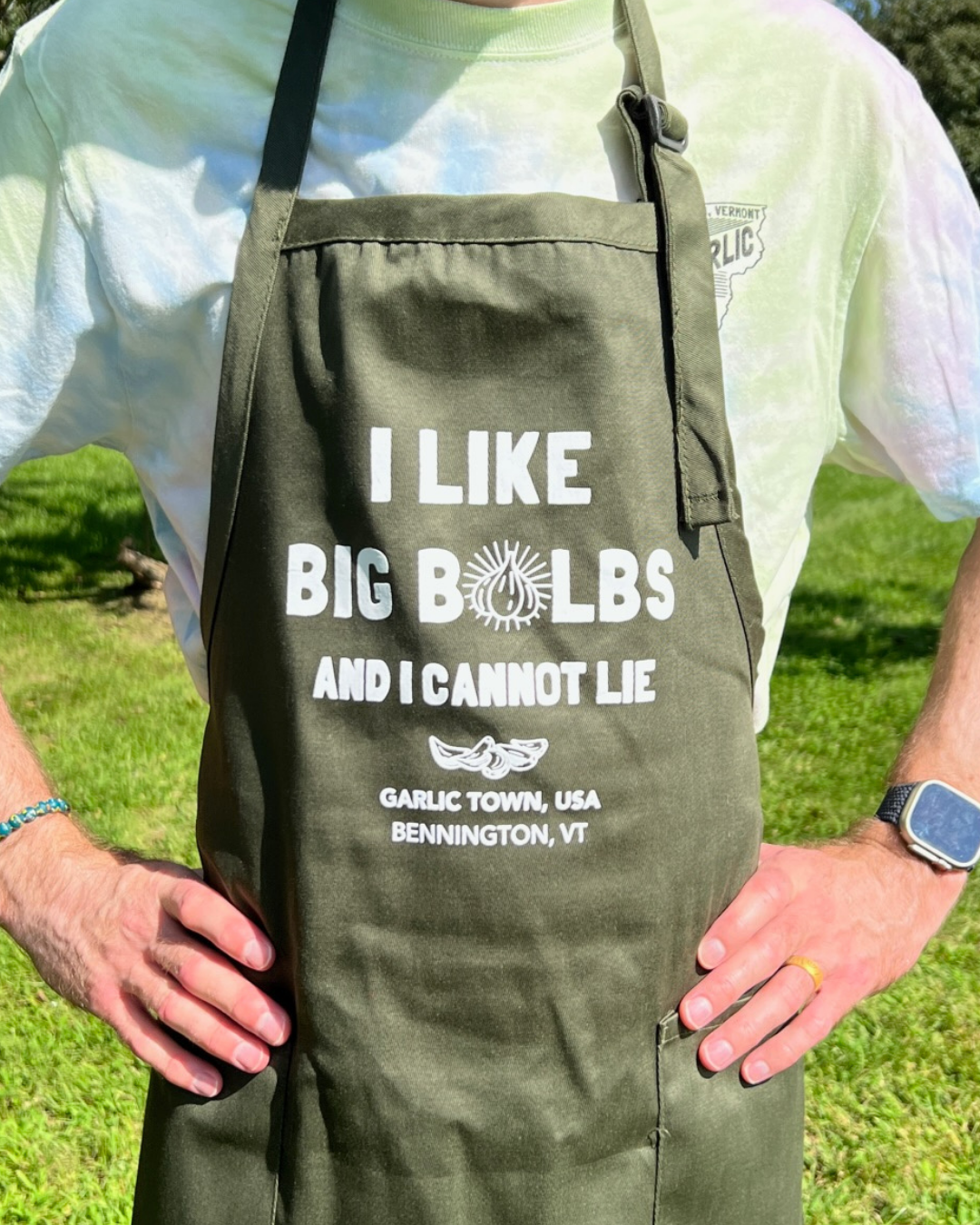 Person wearing a white t-shirt with a patch on the shoulder, carrying a black apron with the text: 'I LIKE BIG BOLBS AND I CANNOT LIE, GARLIC TOWN, USA, BENNINGTON, VT' and an illustration of garlic. The person is outdoors on green grass, with one ha