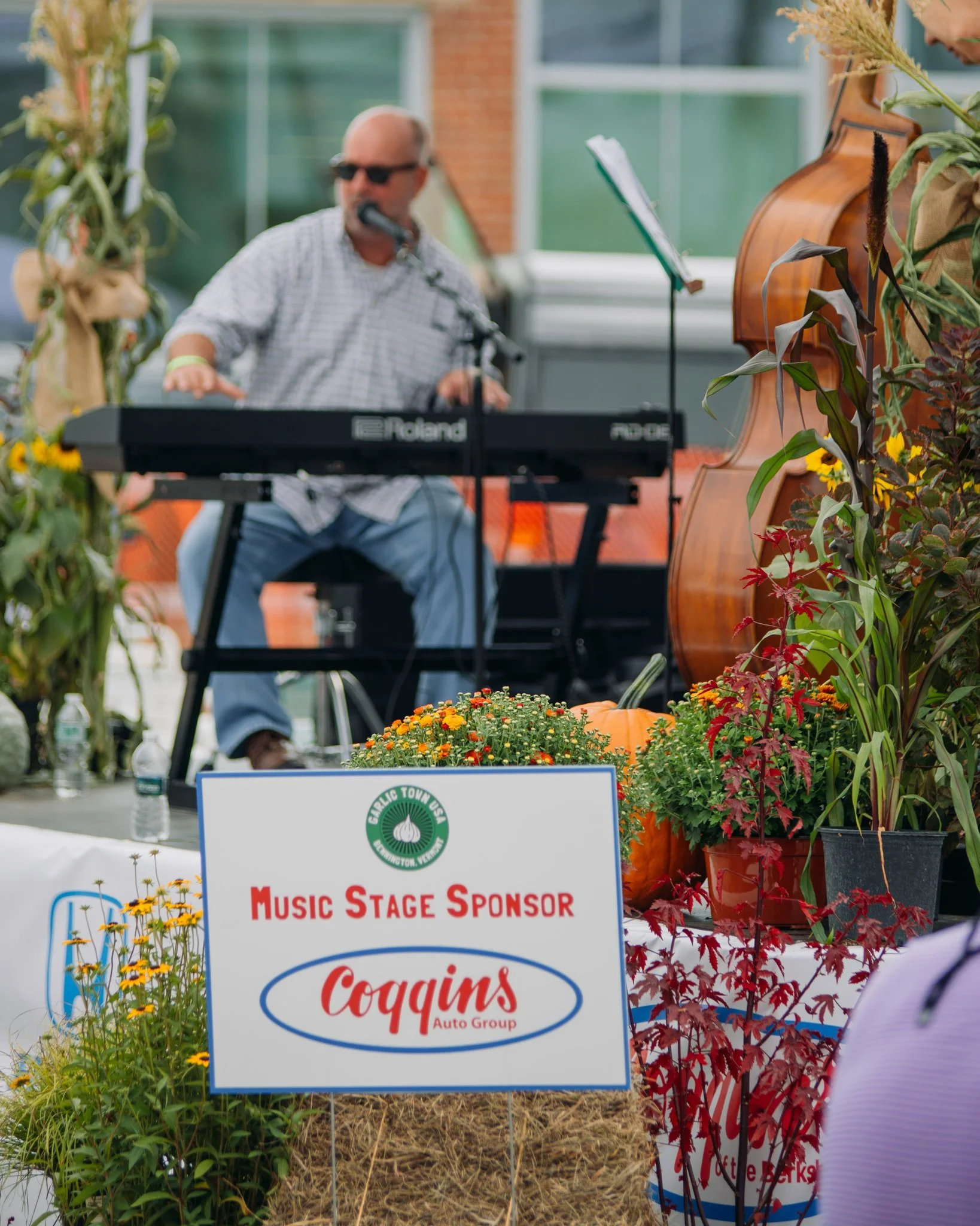 A musician playing a keyboard outdoors, with a double bass and autumn decorations surrounding him. A sign indicates sponsorship from Coqgin's Auto Group.