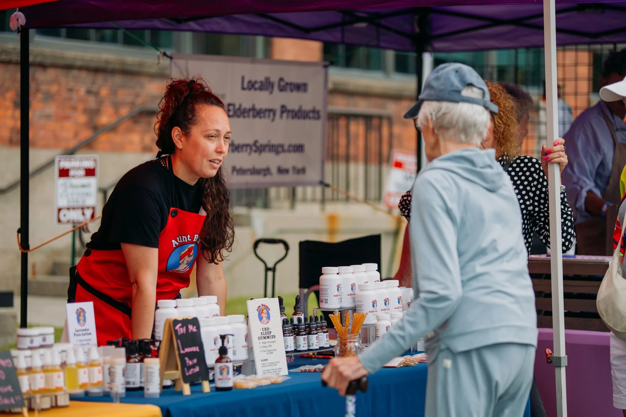 A woman with long curly hair wearing a black shirt and red apron is talking to an elderly woman at an outdoor market stall. The stall displays various health and wellness products like bottles, jars, and gummies and has signs indicating locally grown