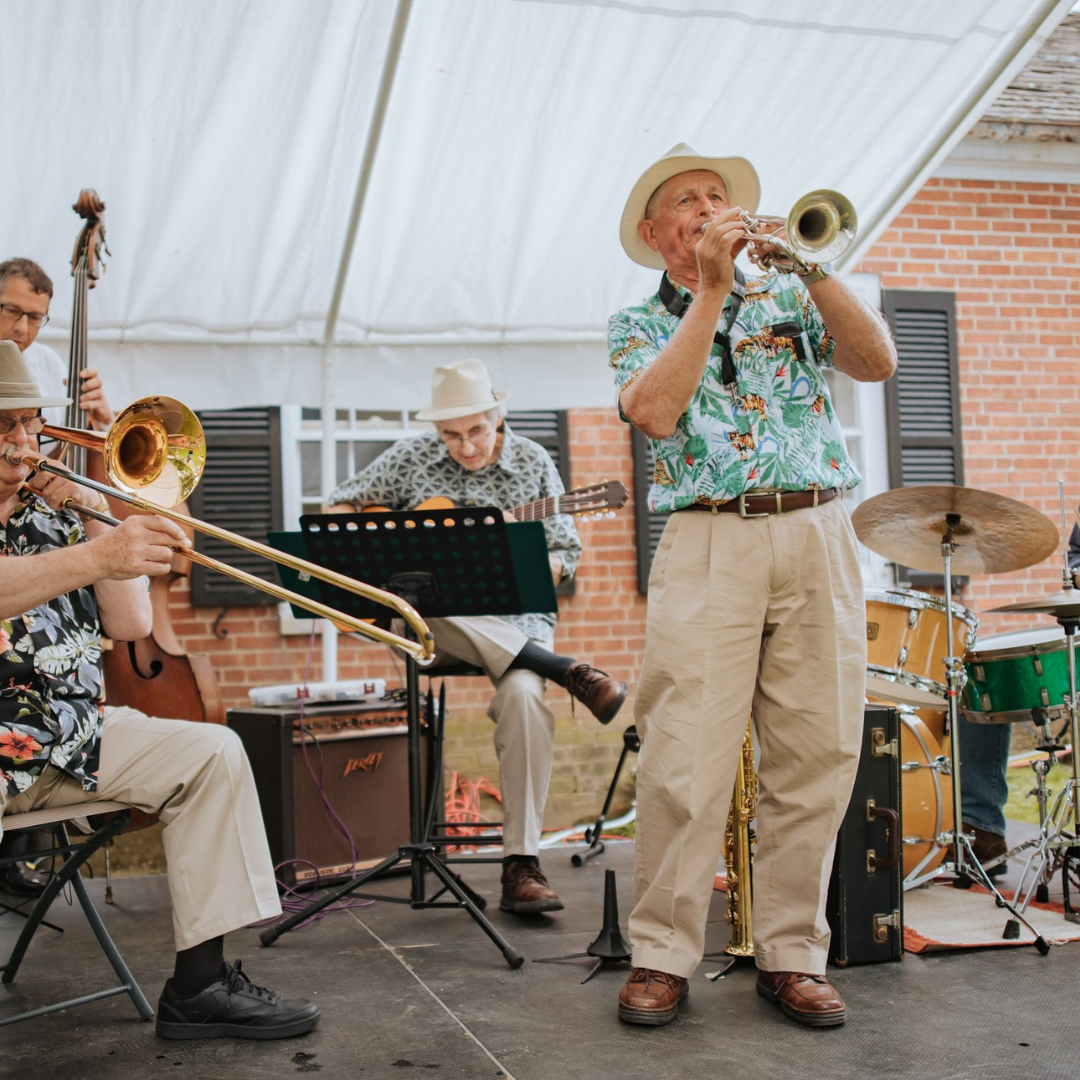 A group of elderly men performing jazz music outdoors, playing various instruments including trumpet, trombone, guitar, and drums, under a white canopy with a brick building in the background.