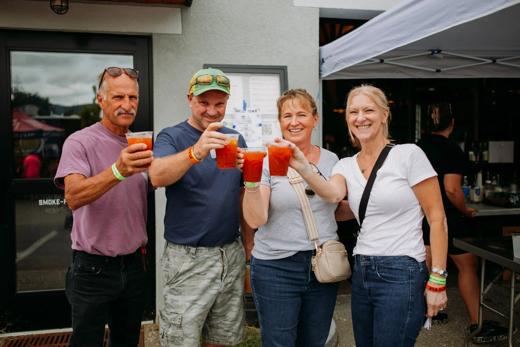 Four adults smiling, holding drinks, standing outside a building with a white canopy overhead, celebrating together.