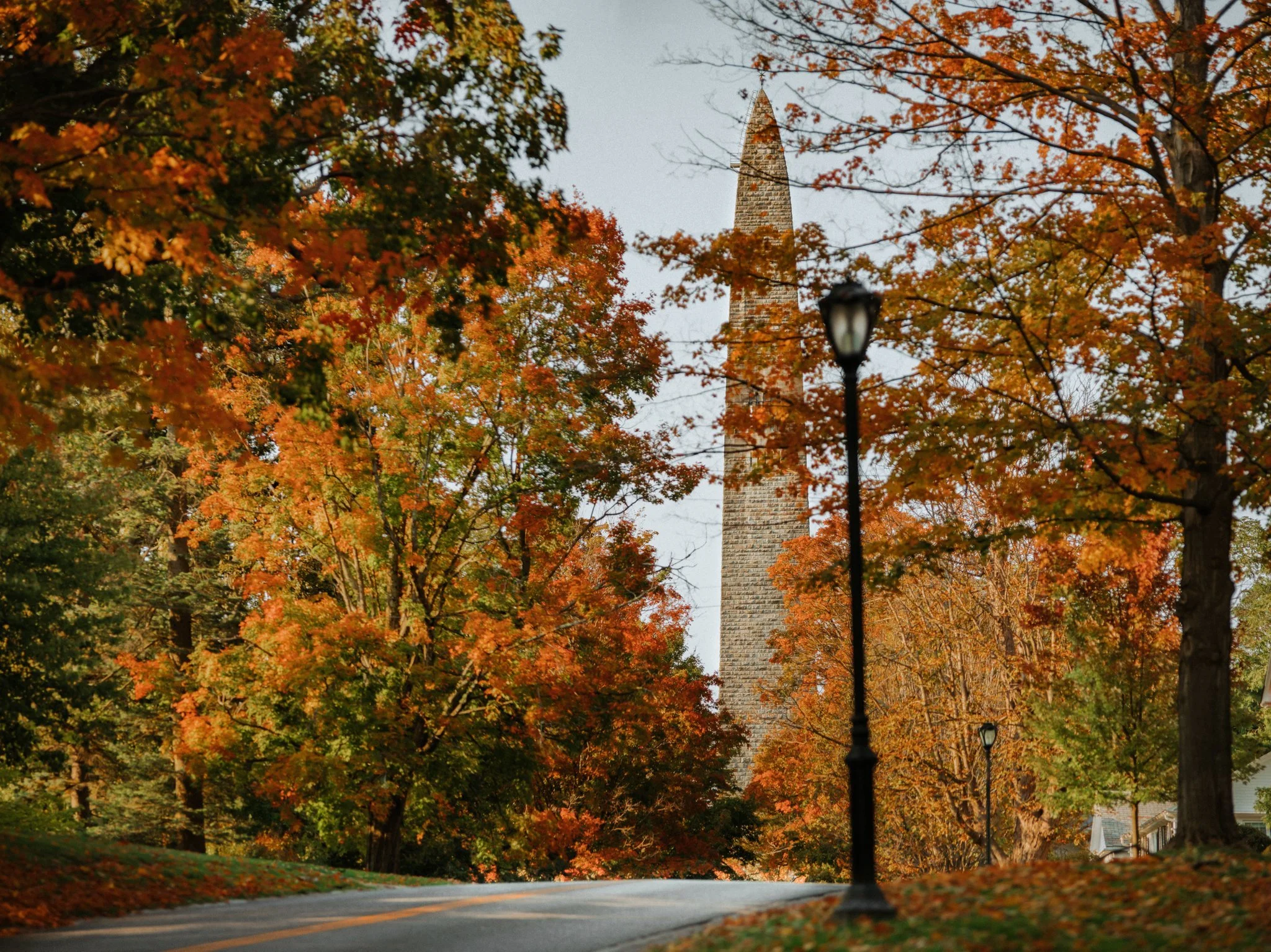A winding road lined with fall foliage trees with orange leaves and a tall stone obelisk in the background, partially obscured by the trees. A black streetlamp is in the foreground.