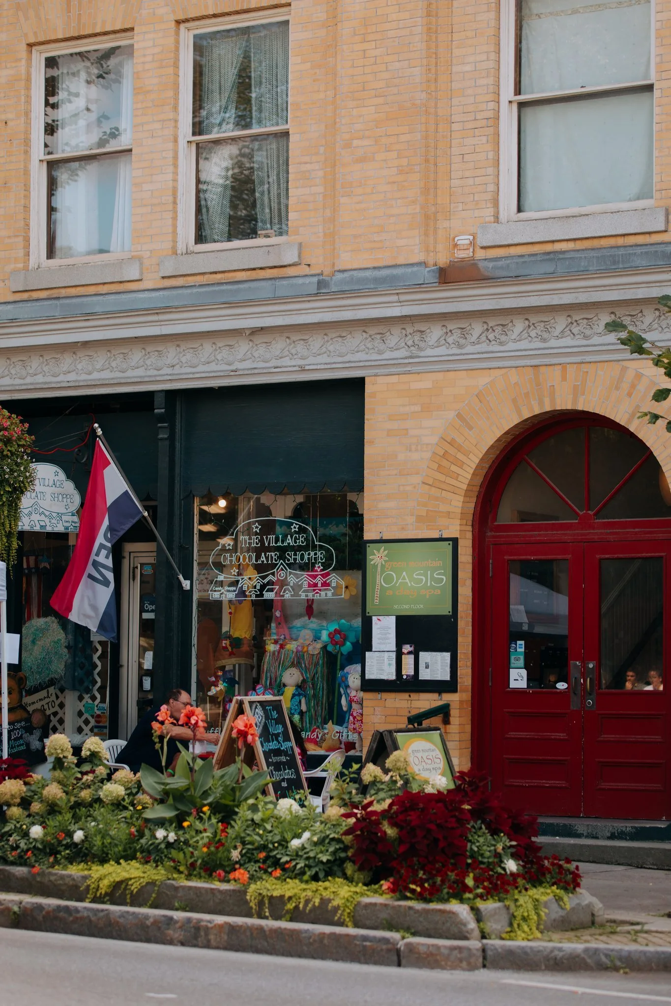 A storefront with a black and yellow brick exterior, two large windows on the upper floor, and a red arched door. The store is called The Village Chocolate Shoppe and is decorated with flowers and signs promoting chocolates, gifts, and a spa called O