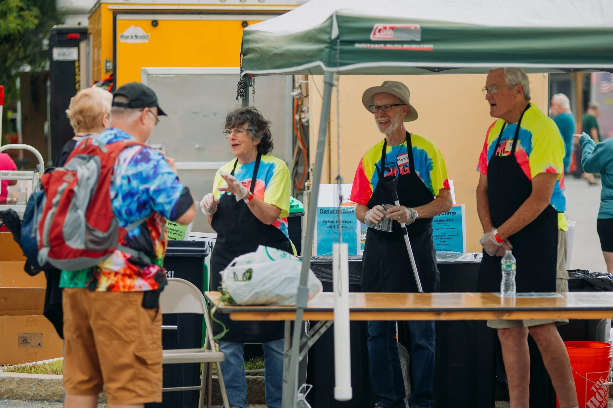 Four volunteers wearing colorful tie-dye shirts and black aprons standing behind a table at an outdoor event, talking to a man with a backpack, under a canopy tent.