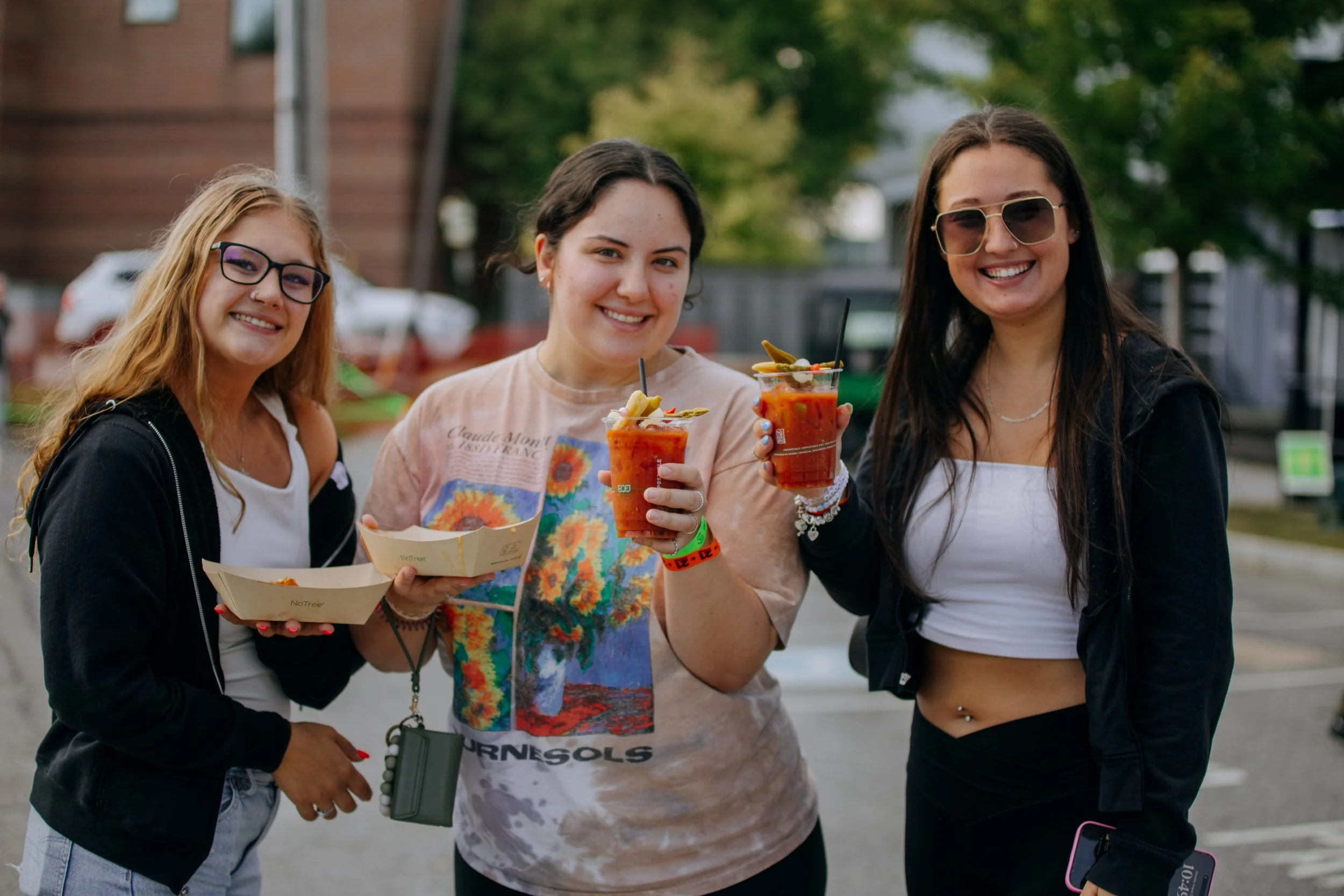 Three young women smiling and holding drinks and food outdoors at a festival or event.