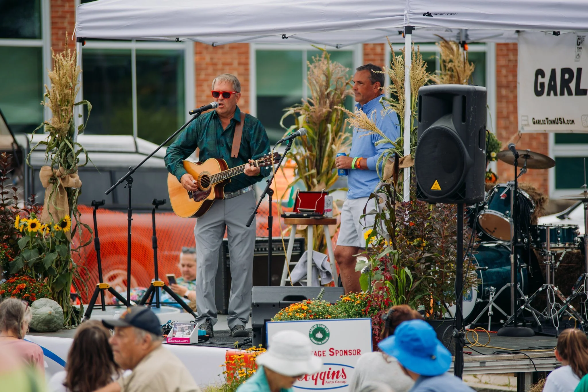 A man playing guitar and singing into a microphone on an outdoor stage, with another man standing beside him holding a water bottle, surrounded by fall decorations, musical equipment, and audience members seated in front.
