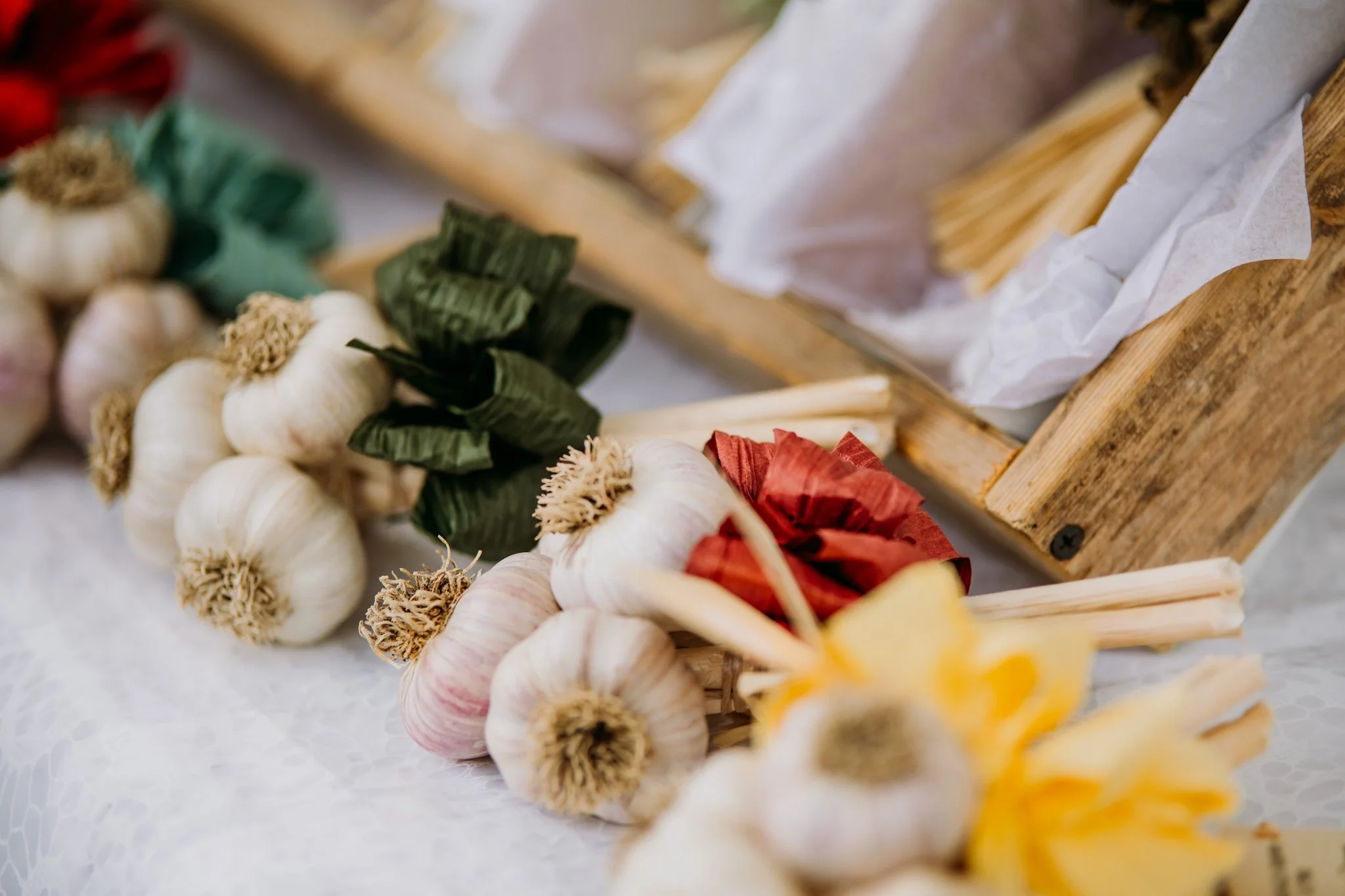 A display of fresh garlic bulbs with their roots and papery skins, arranged on a white tablecloth near a wooden crate.