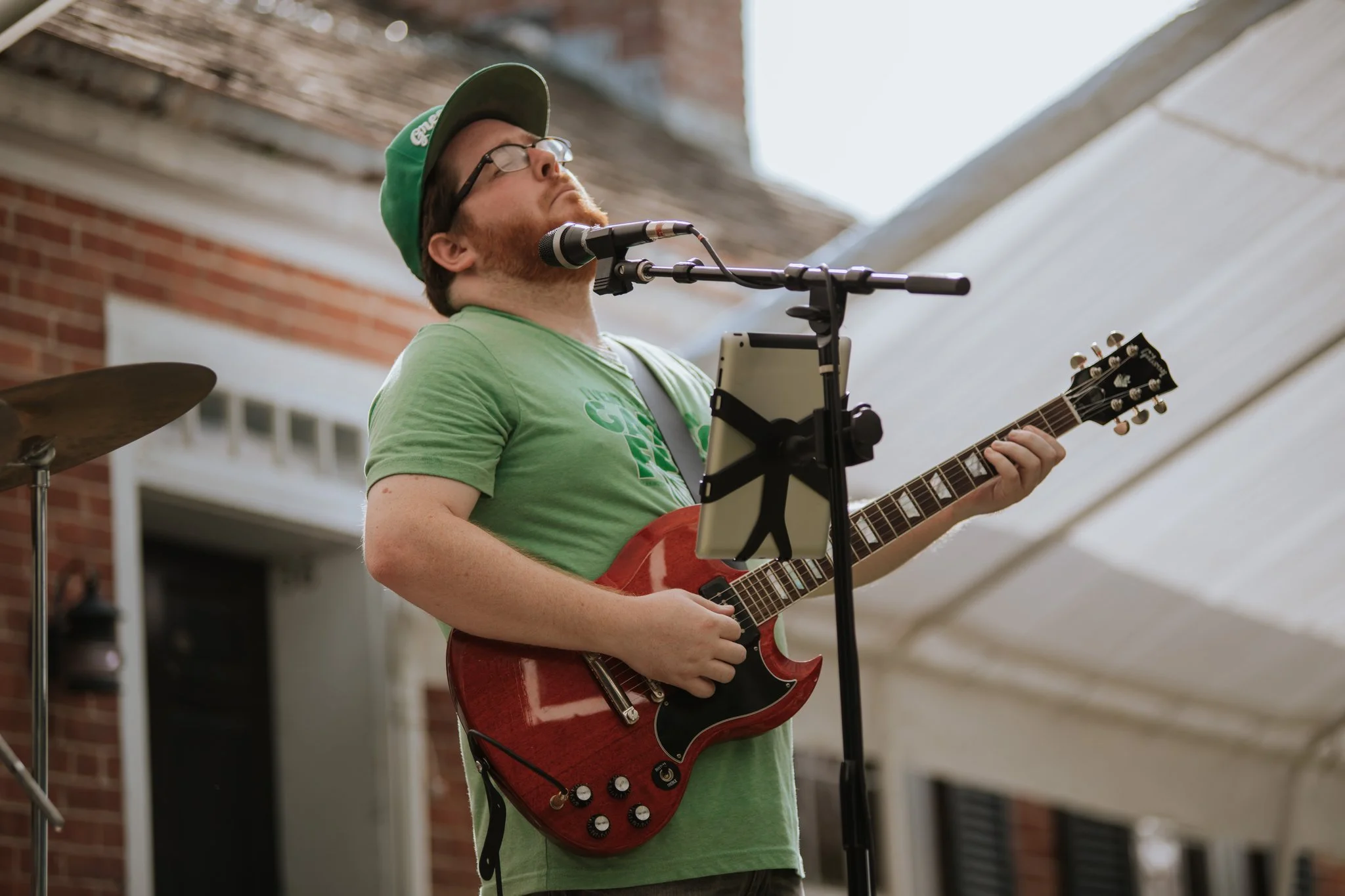 A man wearing glasses, a green cap, and a green t-shirt sings into a microphone while playing a red electric guitar during a live outdoor performance.
