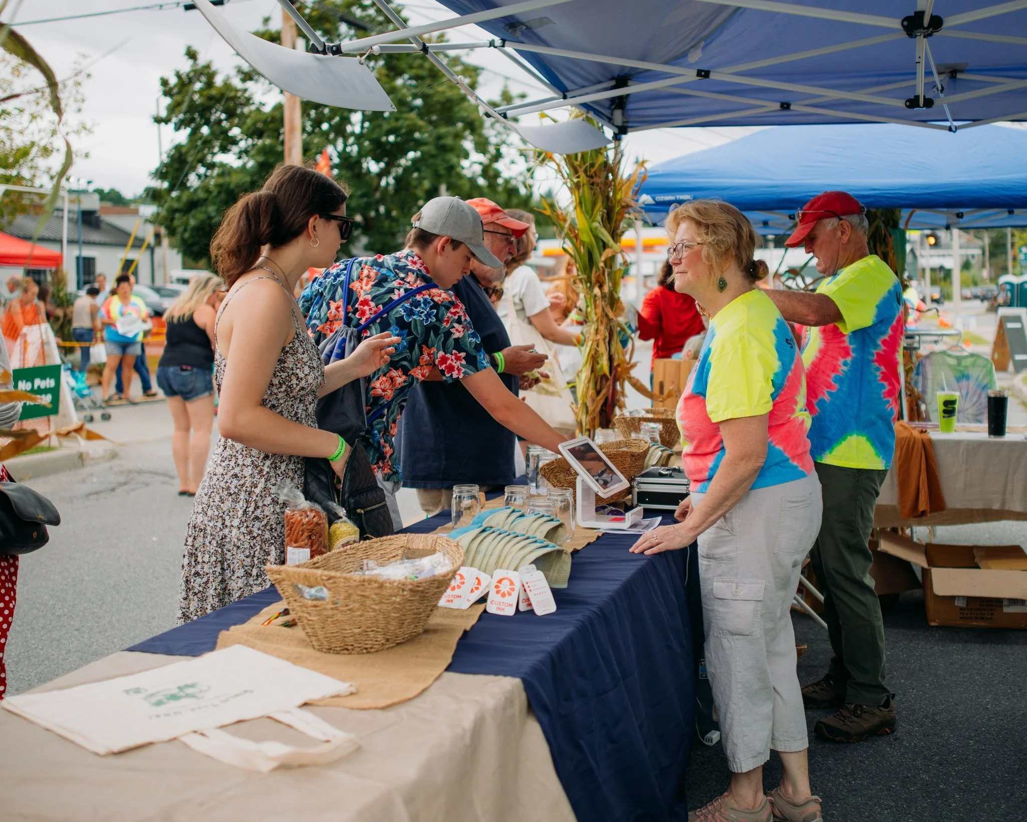 People shopping at an outdoor farmers market stall with various products on display, including jars, bags of snacks, and a digital payment device, under a blue canopy.