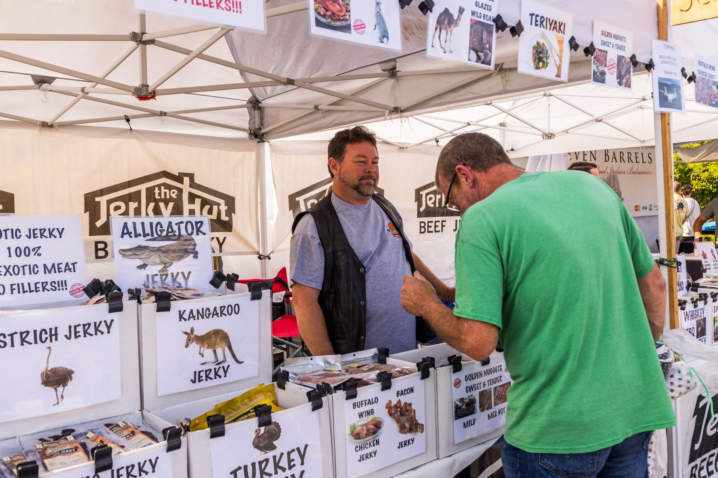 A man in a green shirt shops at a booth selling jerky at an outdoor market. The booth has signs advertising exotic jerky, including alligator, kangaroo, ostrich, and buffalo. The vendor, wearing a vest, interacts with the customer, who is looking at 