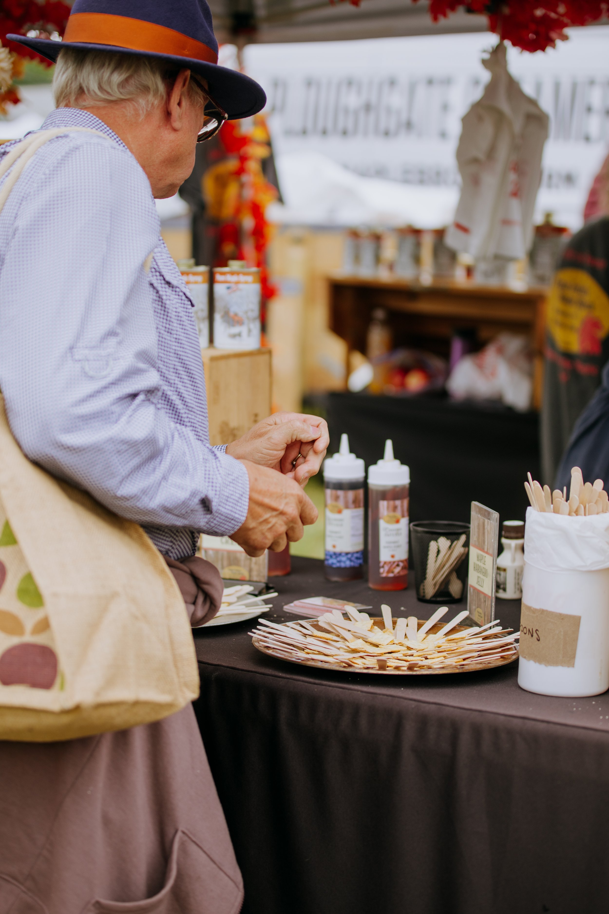 An elderly man with blond hair, wearing a dark blue hat, glasses, a light purple shirt, and a beige apron, standing at an outdoor food stall. The stall has bottles of sauces, a plate of wooden tasting sticks, and containers with smiling faces. The ba